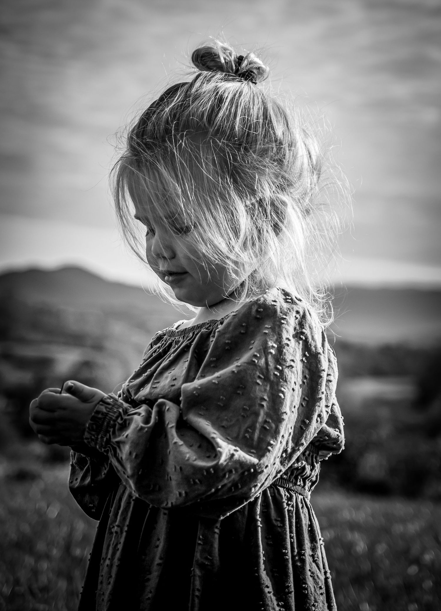 Black and white portrait of a child in vintage dress looking down while standing in a field.