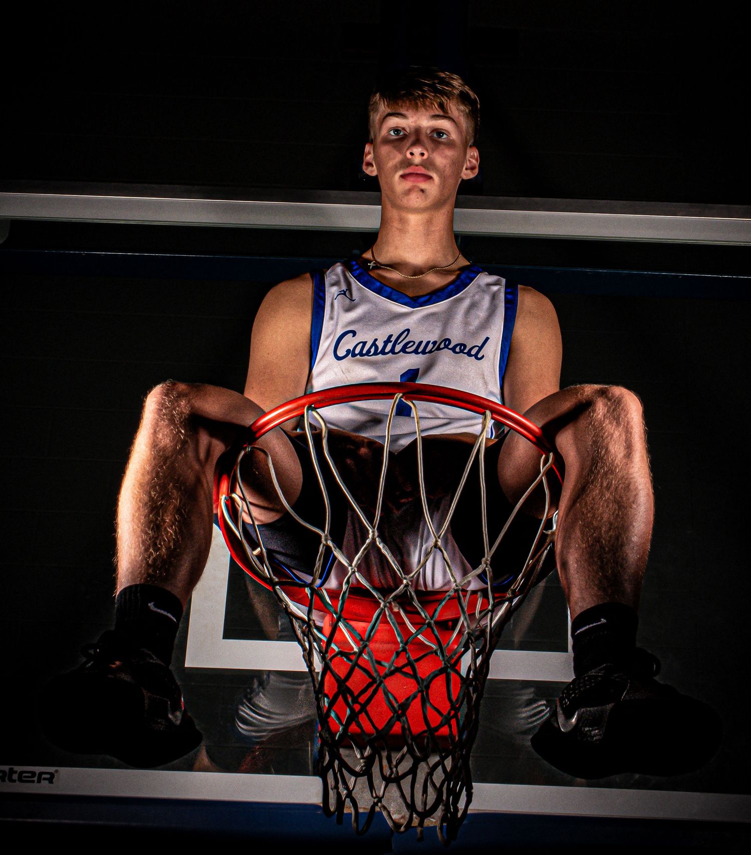 Basketball player sits atop hoop rim in dramatic lighting wearing Castlewood jersey.