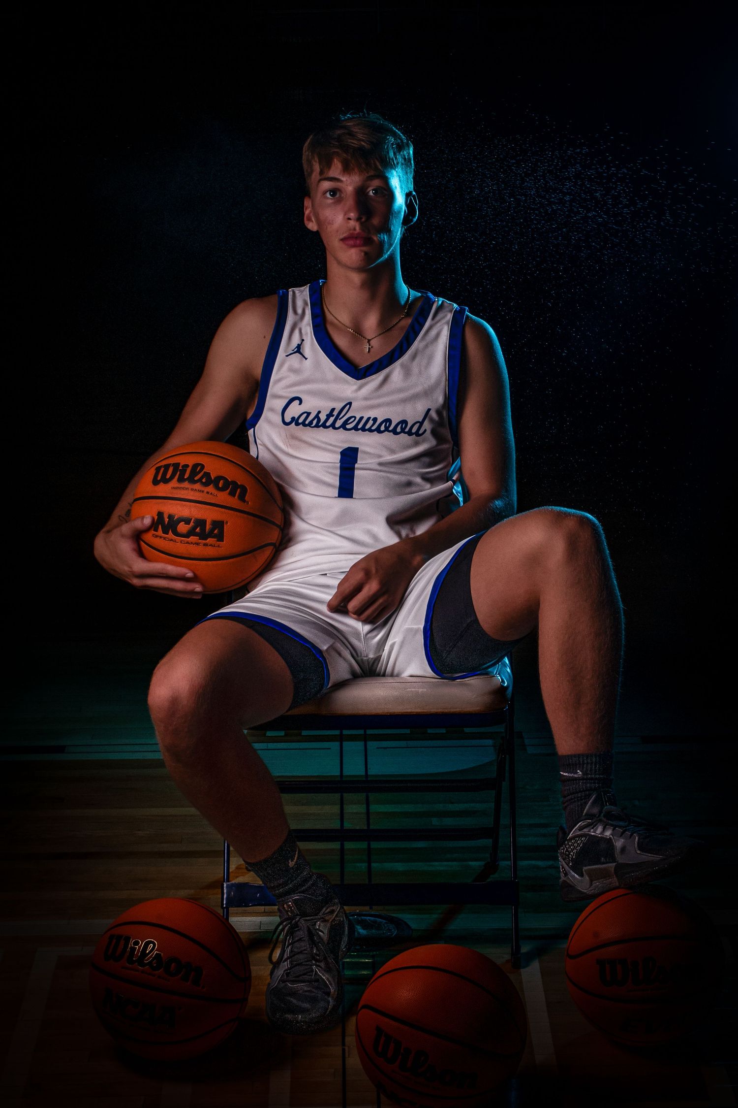 Basketball player in white Castlewood jersey sits with multiple basketballs in dramatic lighting.