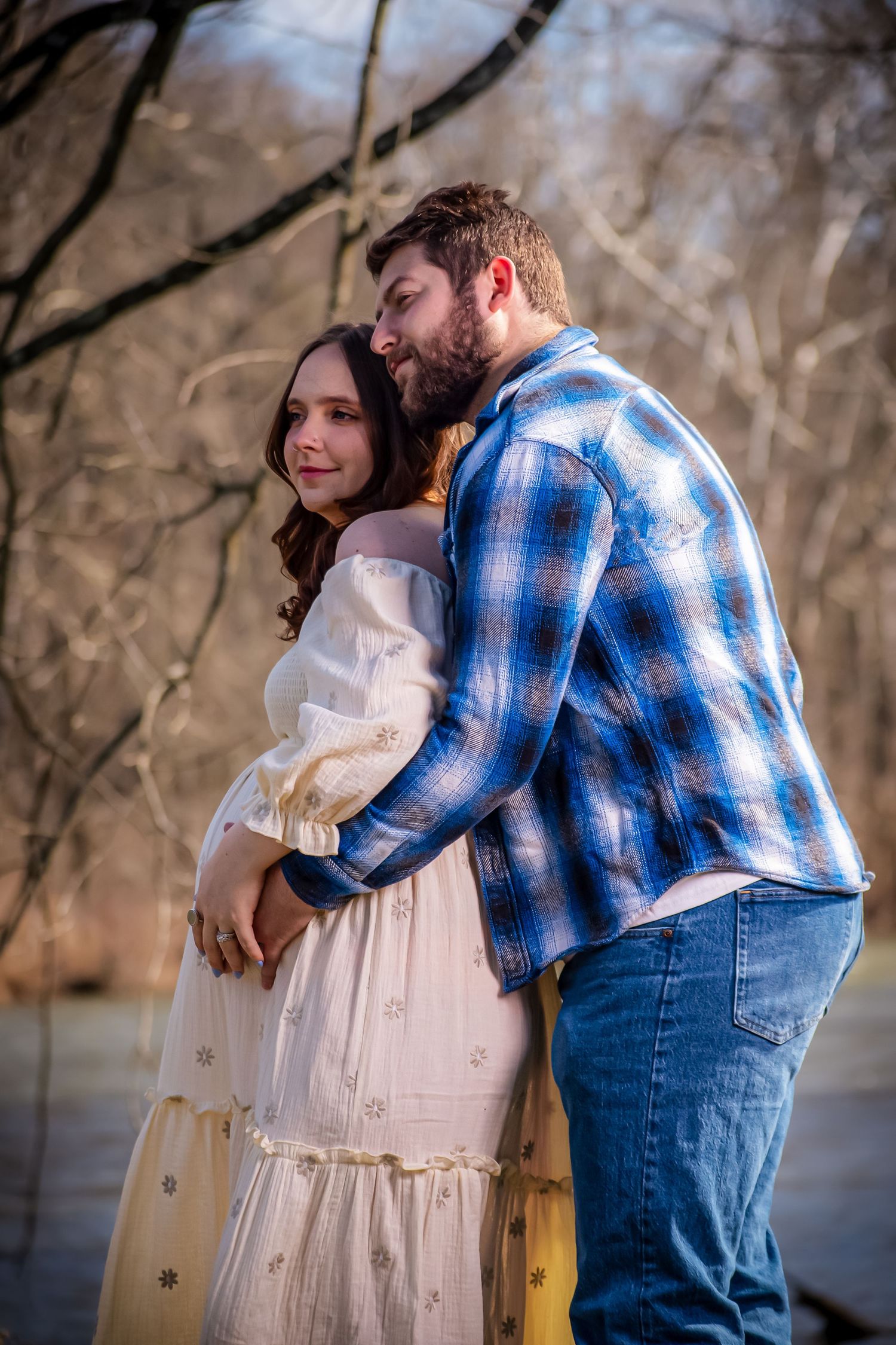 A couple embraces outdoors in a romantic moment, with one wearing a white dress and the other in a blue plaid shirt.