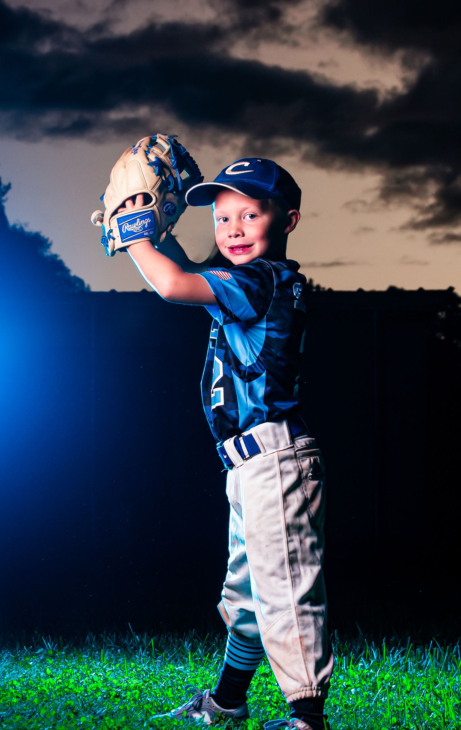 A young baseball player in uniform practices their batting stance against a dramatic stormy sky at dusk.