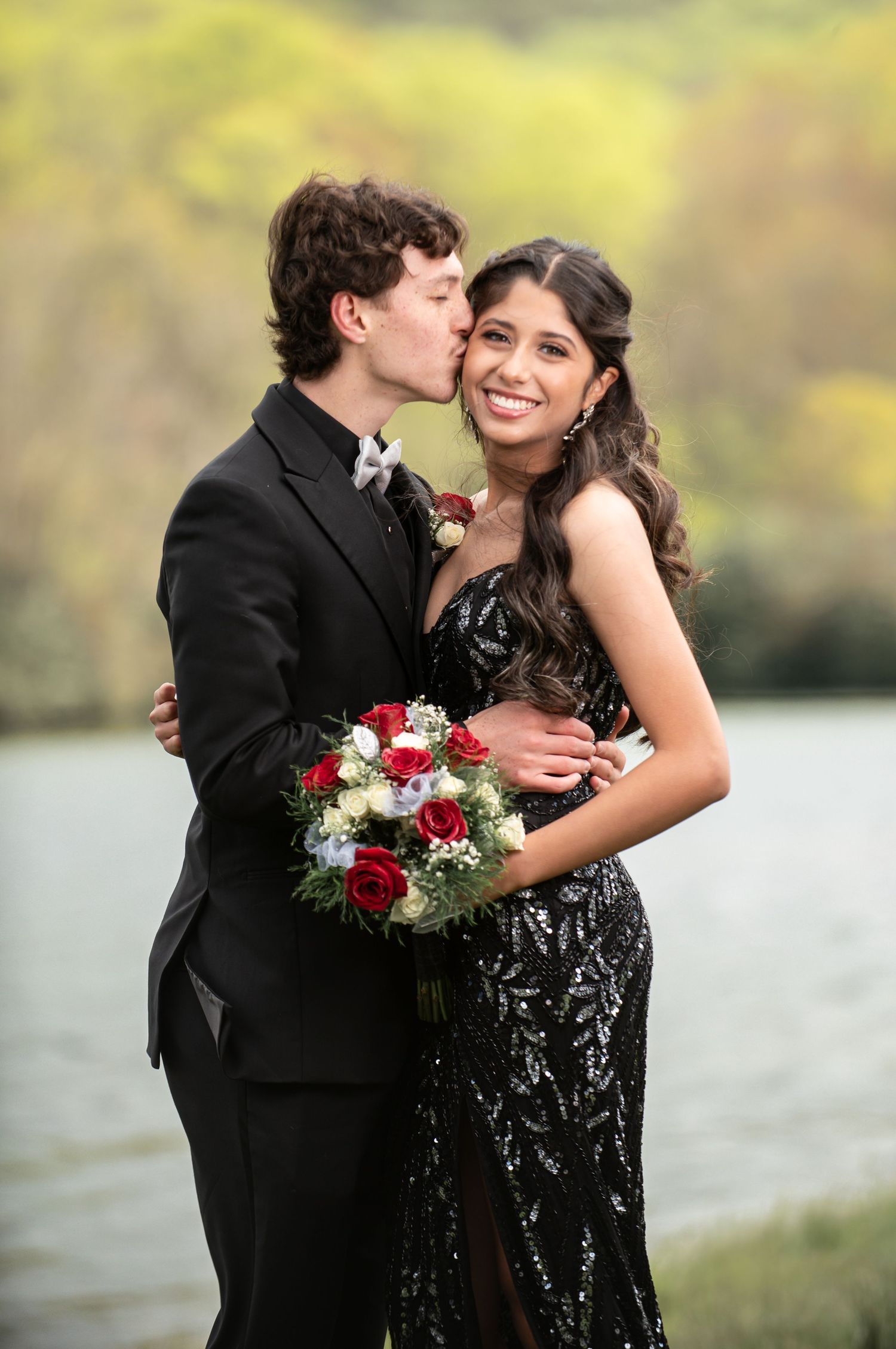 A smiling couple in formal prom attire poses outdoors, the young man kissing her cheek as she holds red roses.