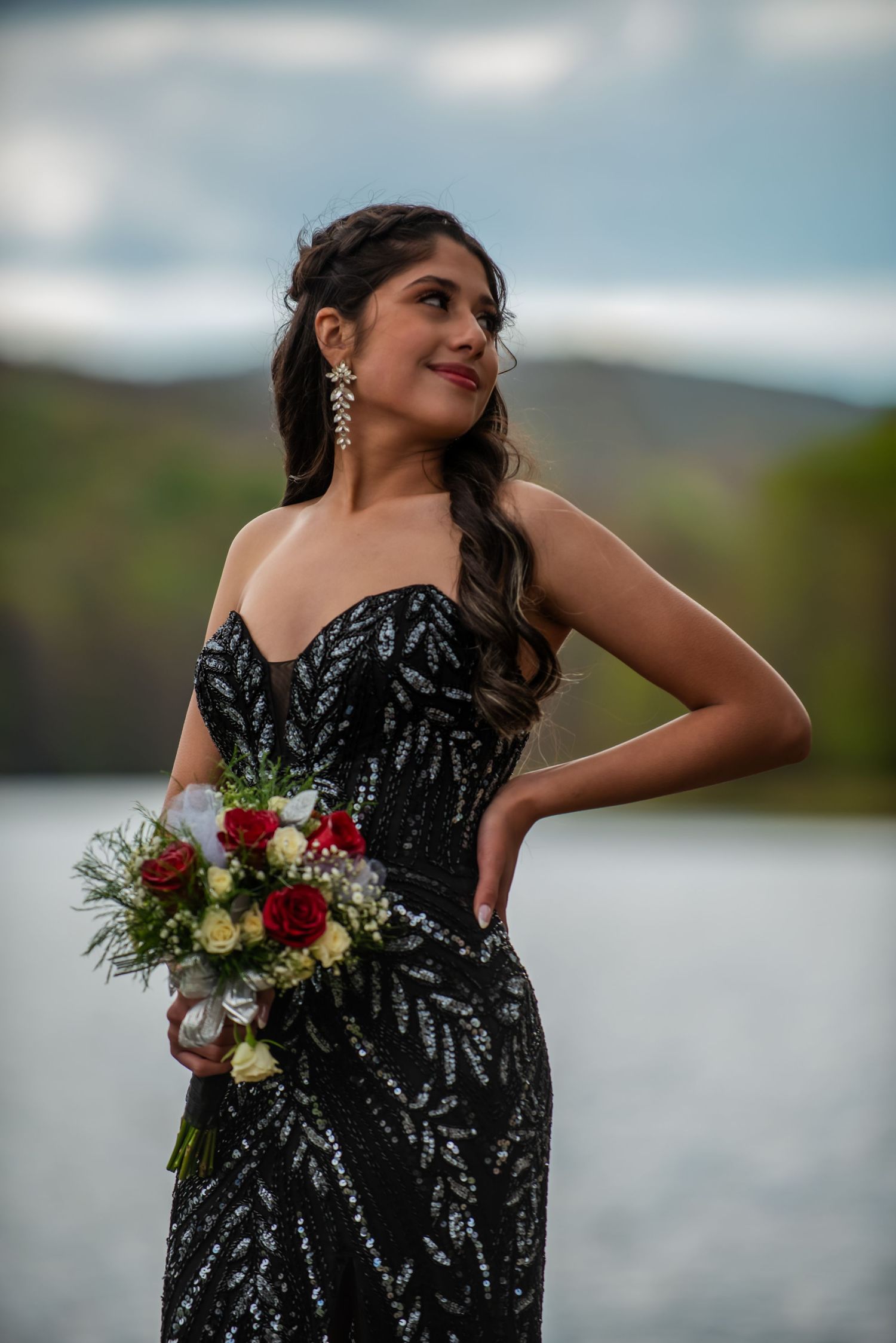 Young woman in black sequined strapless gown holding red and white bouquet, posing outdoors near a lake.