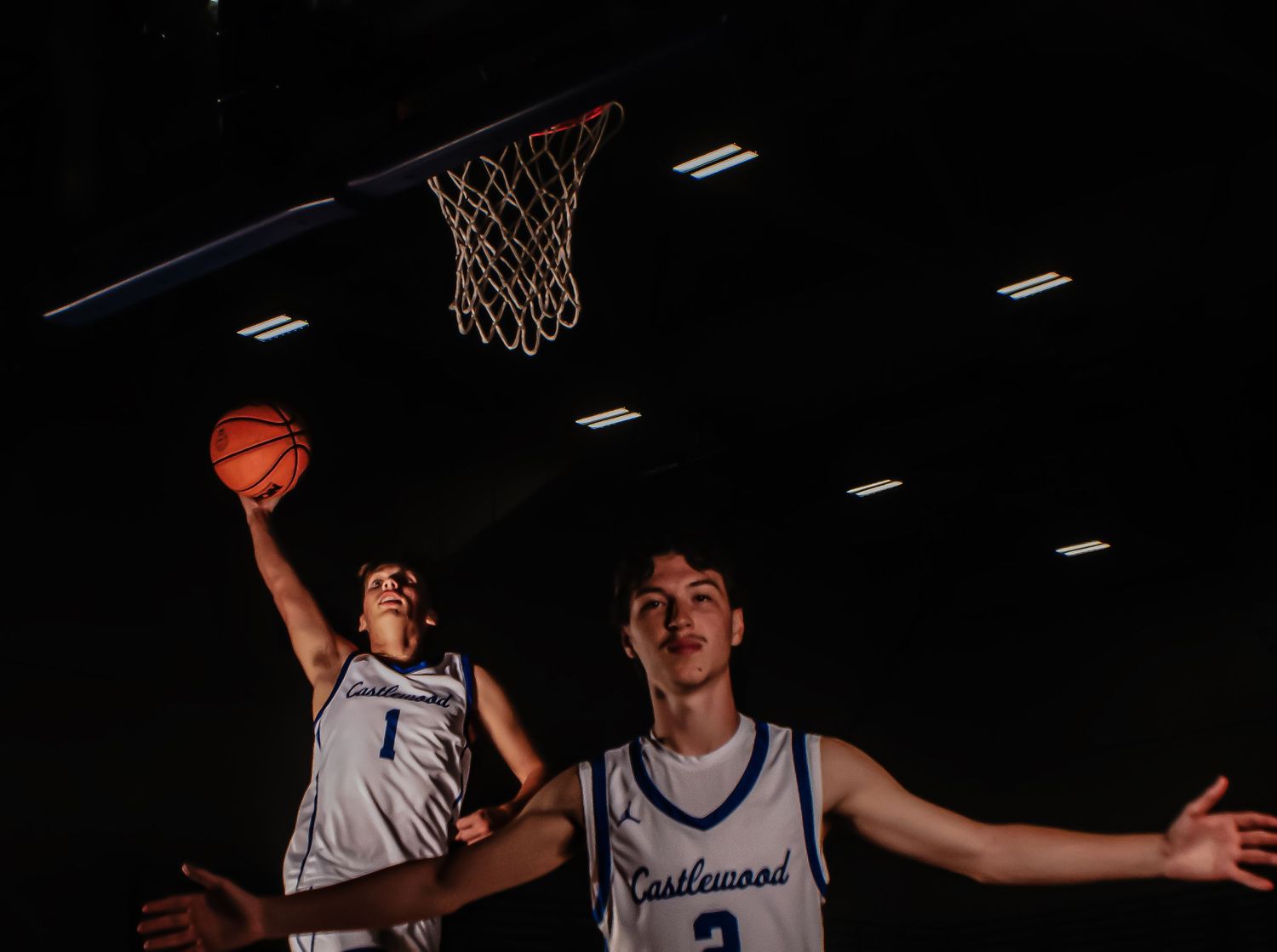 Basketball players reach for ball under illuminated hoop during practice.