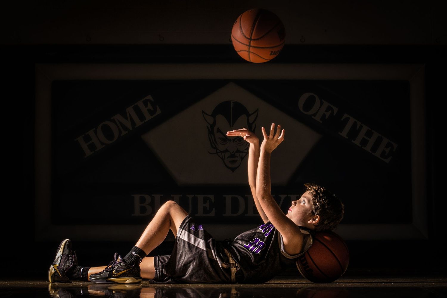 Basketball player lying on court catching a ball in dramatic lighting with 'Home of the' text visible above.