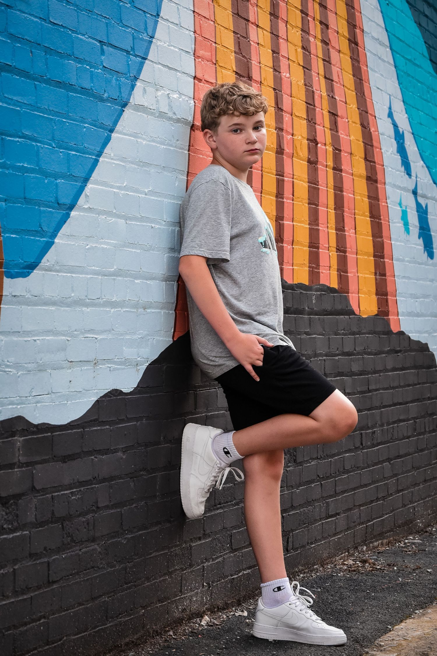 Youth posing against colorful striped mural wall wearing grey t-shirt and shorts.