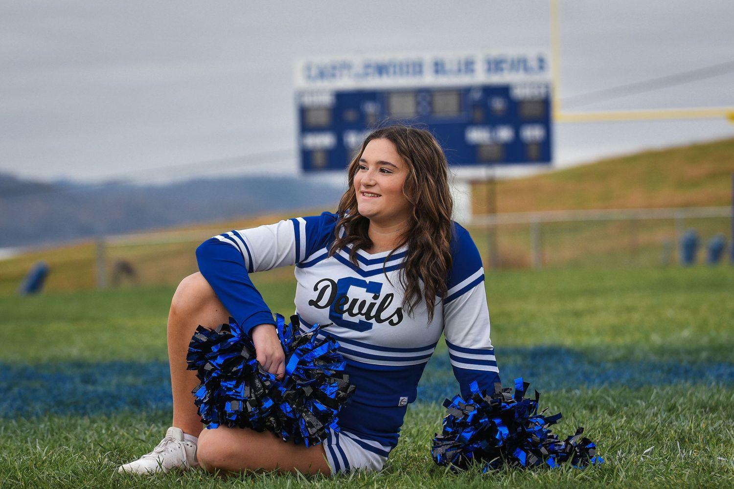Cheerleader in blue and white uniform poses with pom poms on a grassy hillside.