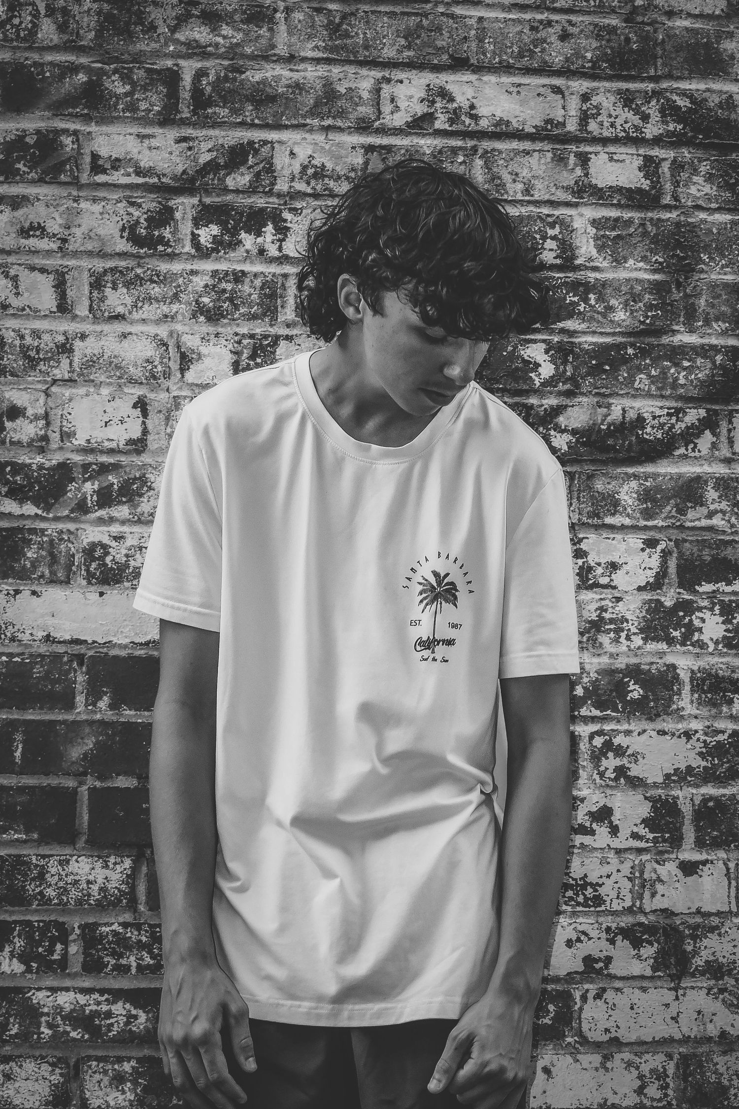 Black and white portrait of person in white t-shirt with small graphic against brick backdrop.