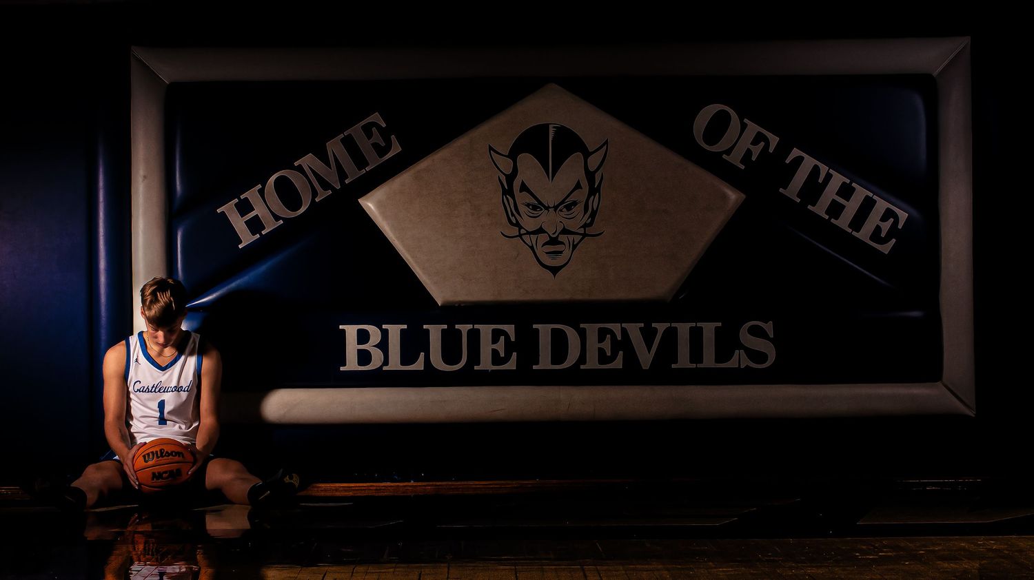 Basketball player in white uniform poses beneath dramatic Blue Devils signage.