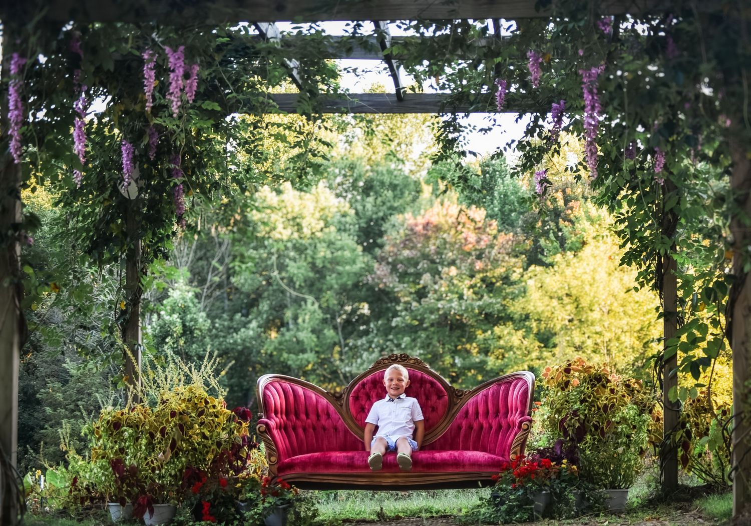 Red velvet couch sits under a wooden pergola in a lush garden setting.