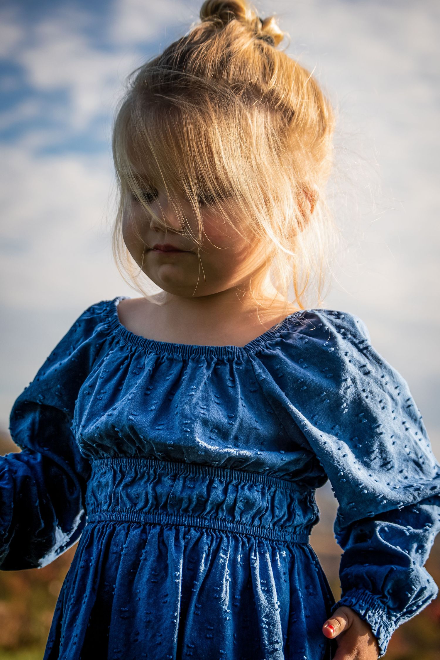 Close-up of child wearing blue smocked dress with gathered sleeves against cloudy sky.