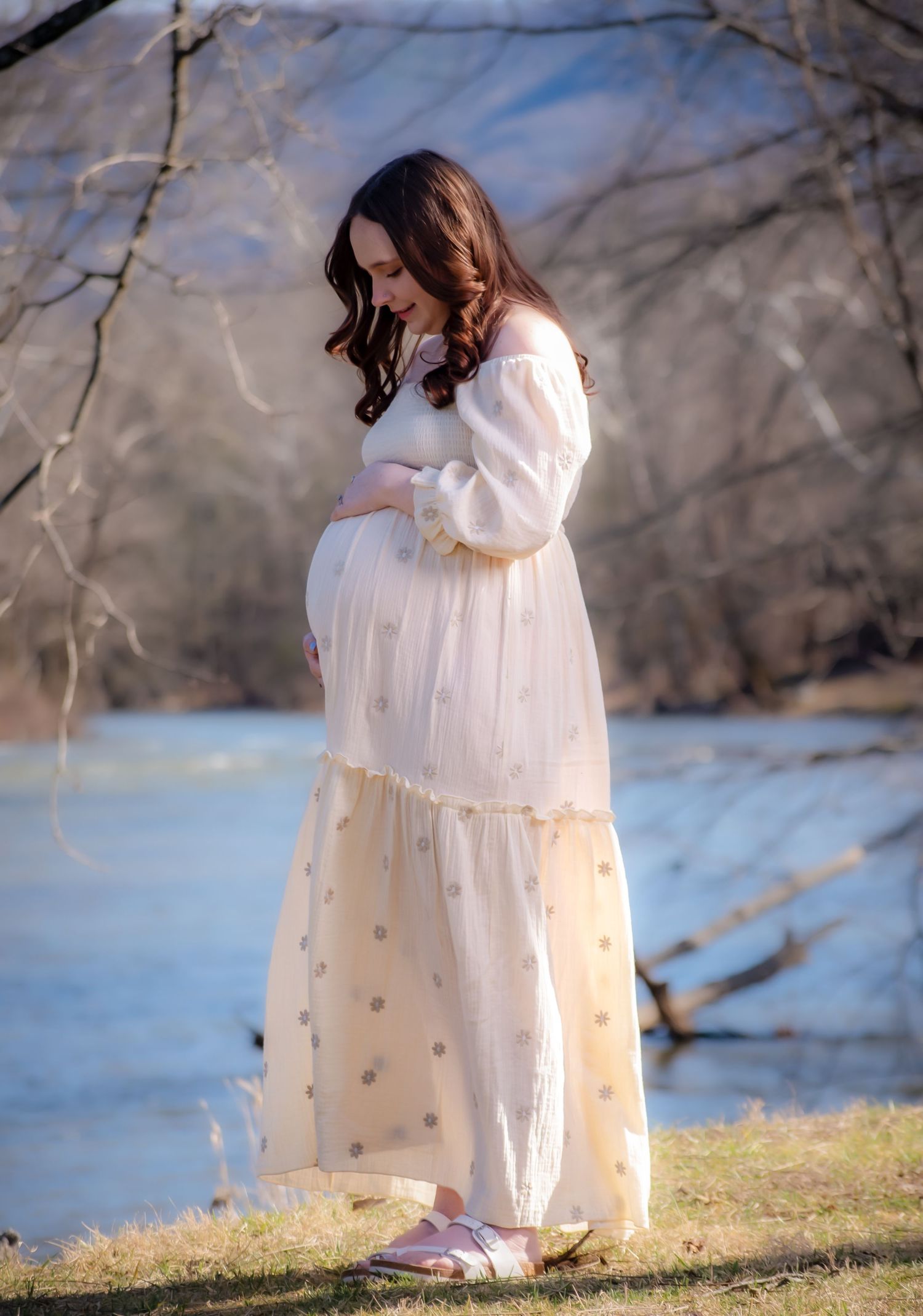 A pregnant woman in a flowing white maxi dress stands by a lake on a winter day, gently cradling her baby bump.