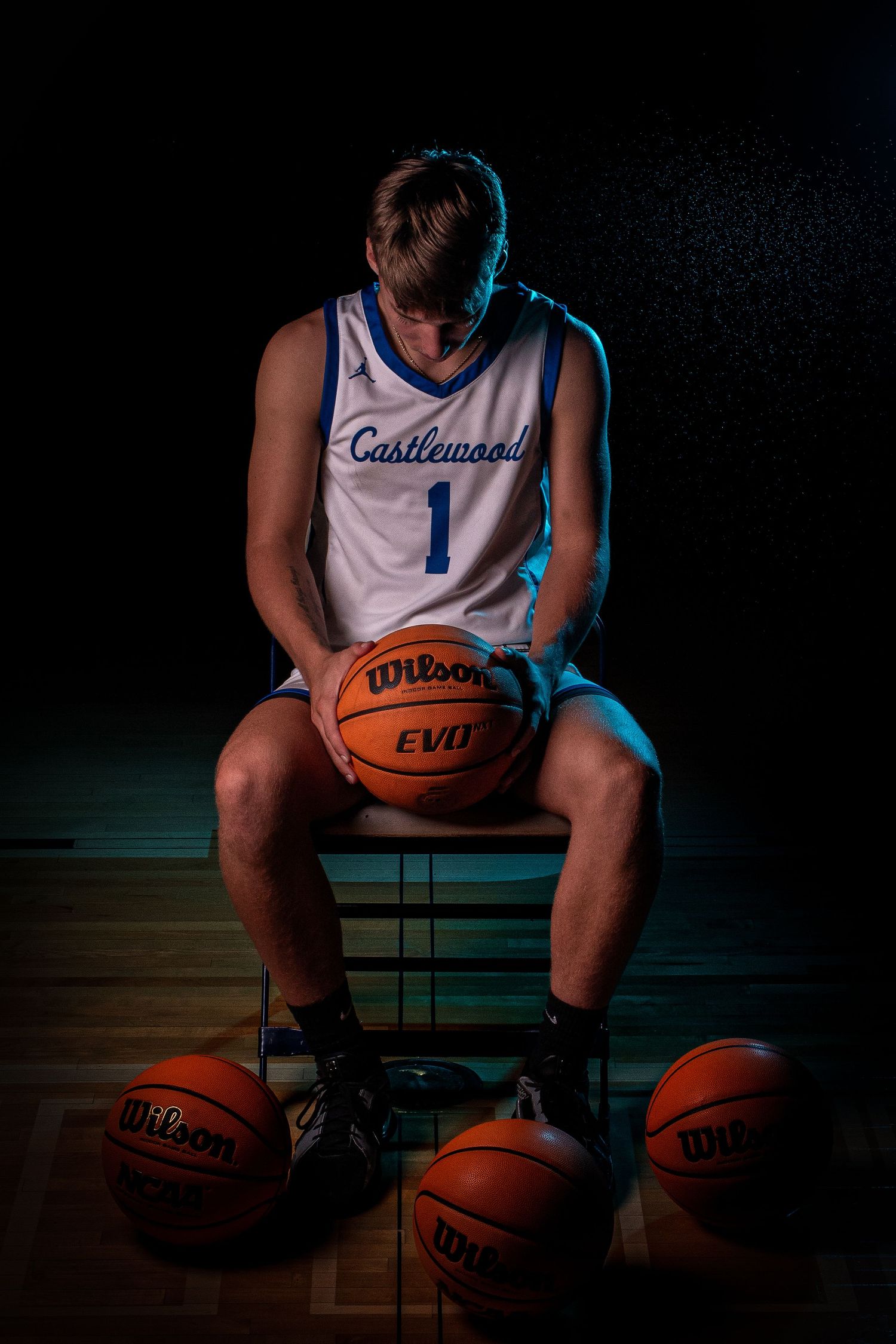 Basketball player sits with multiple Wilson basketballs in dark studio setting.