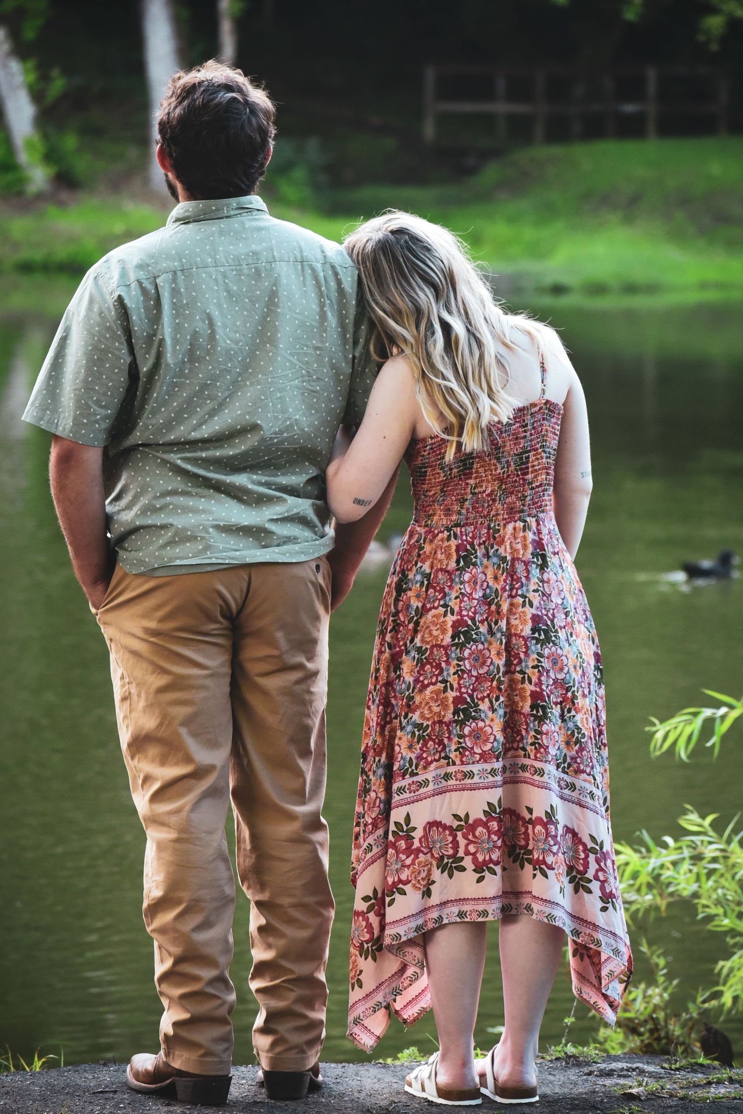 A couple stands side by side on the shore of a peaceful lake during golden hour.