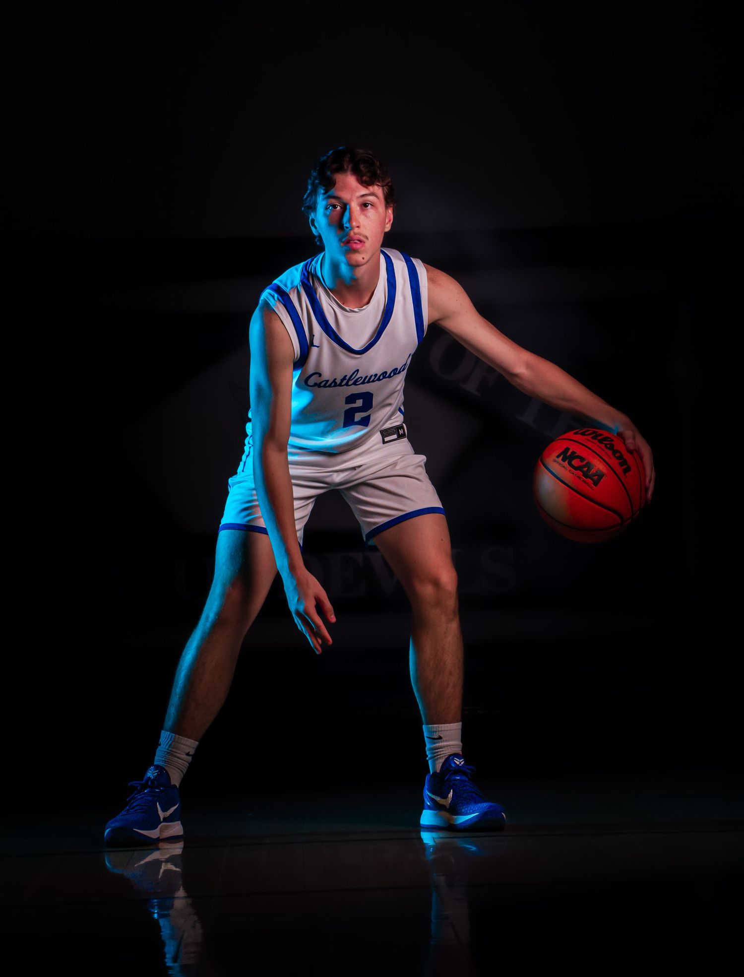 Basketball player in white jersey demonstrates defensive stance with ball under blue lighting.