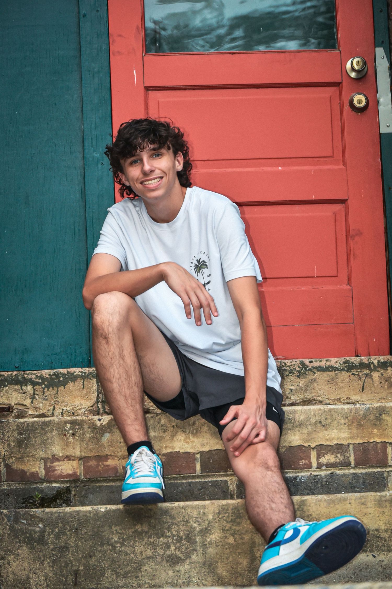 Person in white t-shirt and shorts sits on steps near coral-colored door.