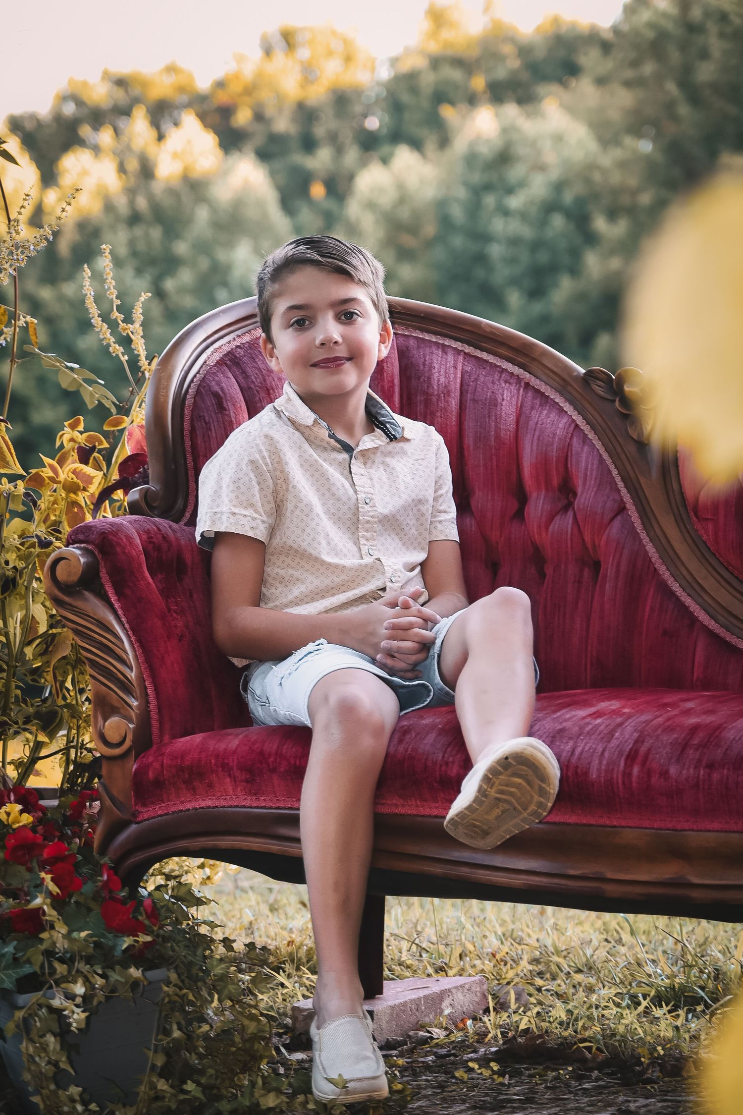 Child sitting on vintage red velvet sofa in white outfit during outdoor photo session at sunset.