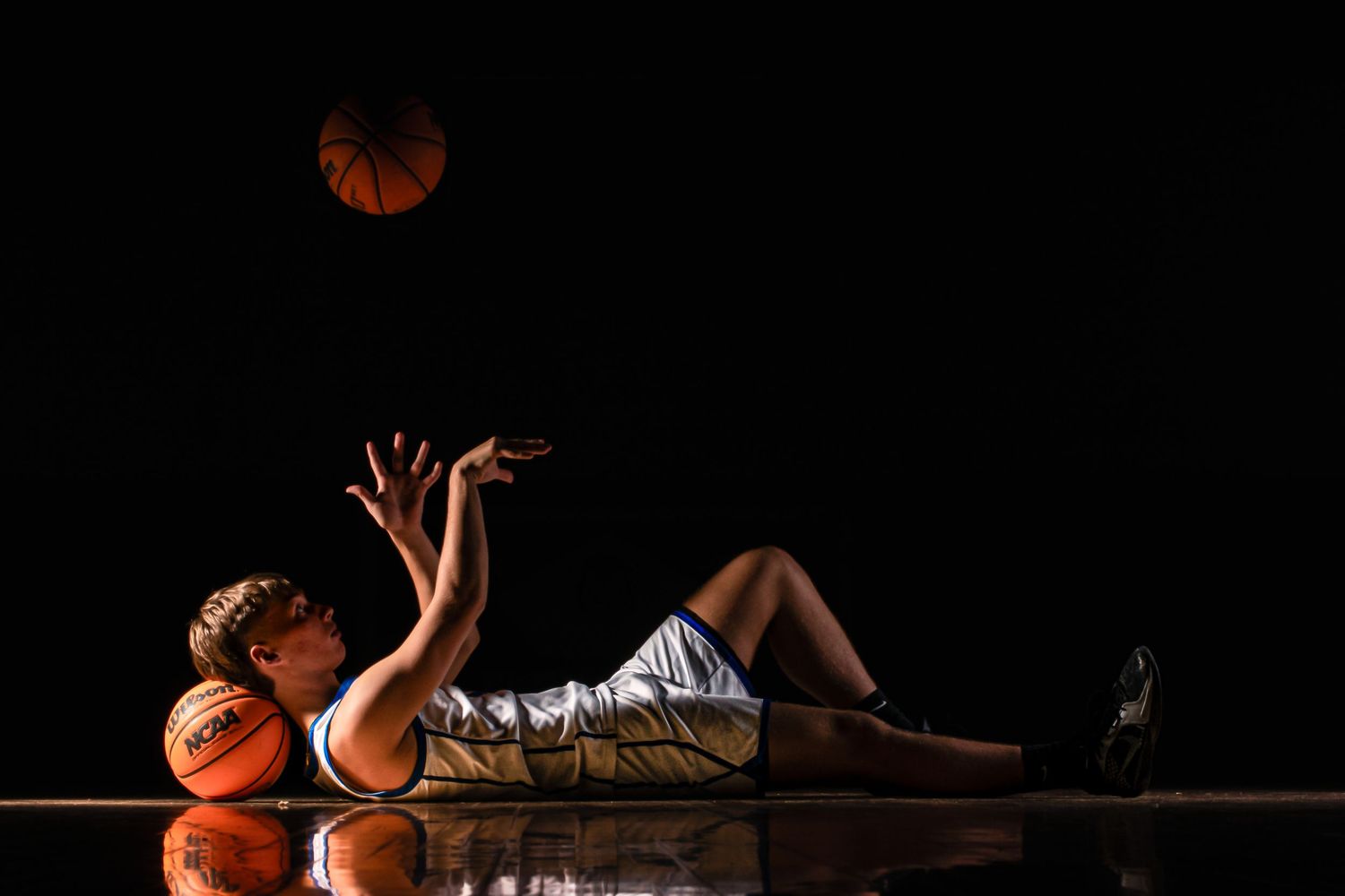 Athlete performs dynamic basketball toss while lying on hardwood court.