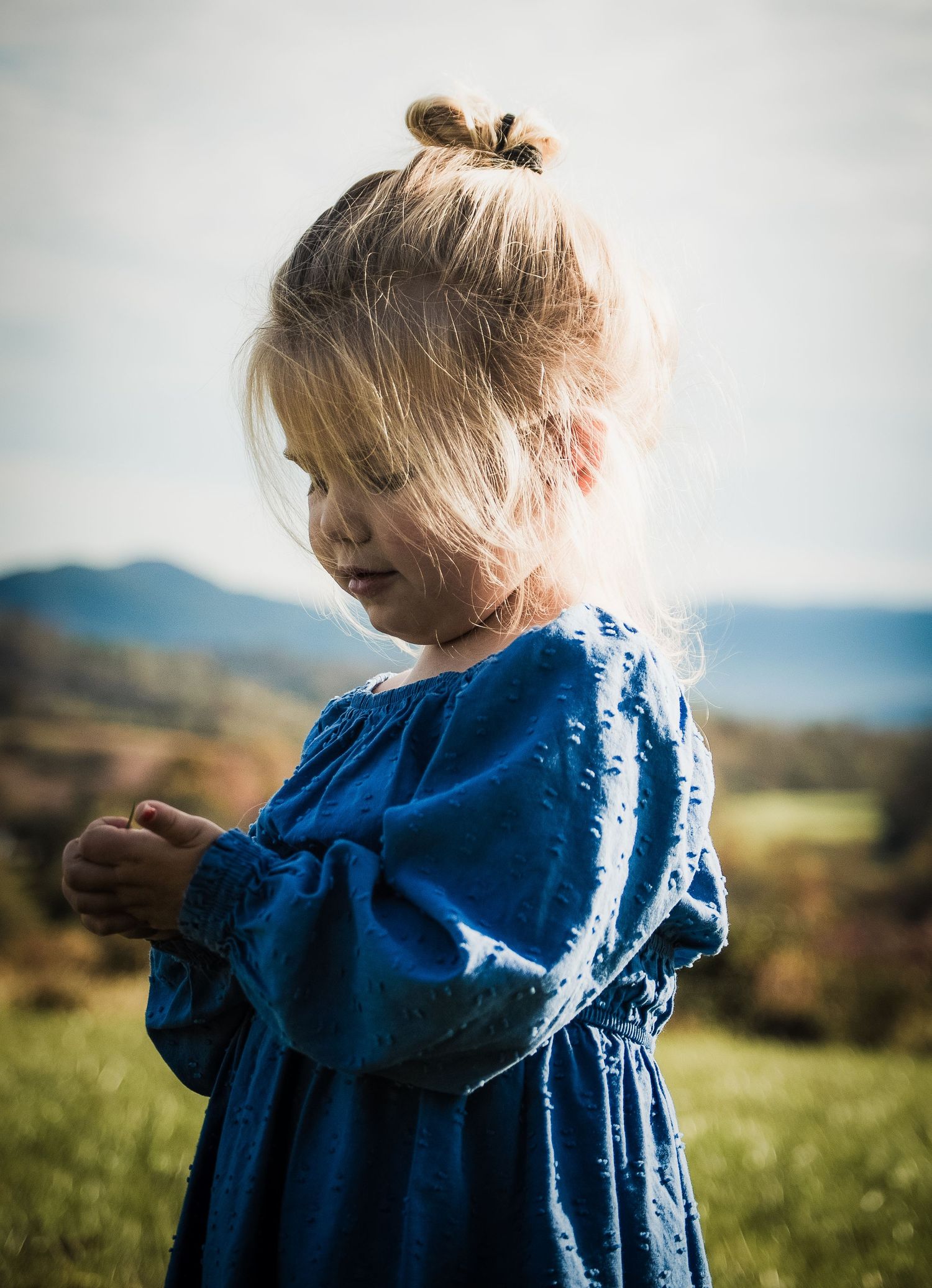 Young child in blue peasant dress examining something while standing in grassy field.