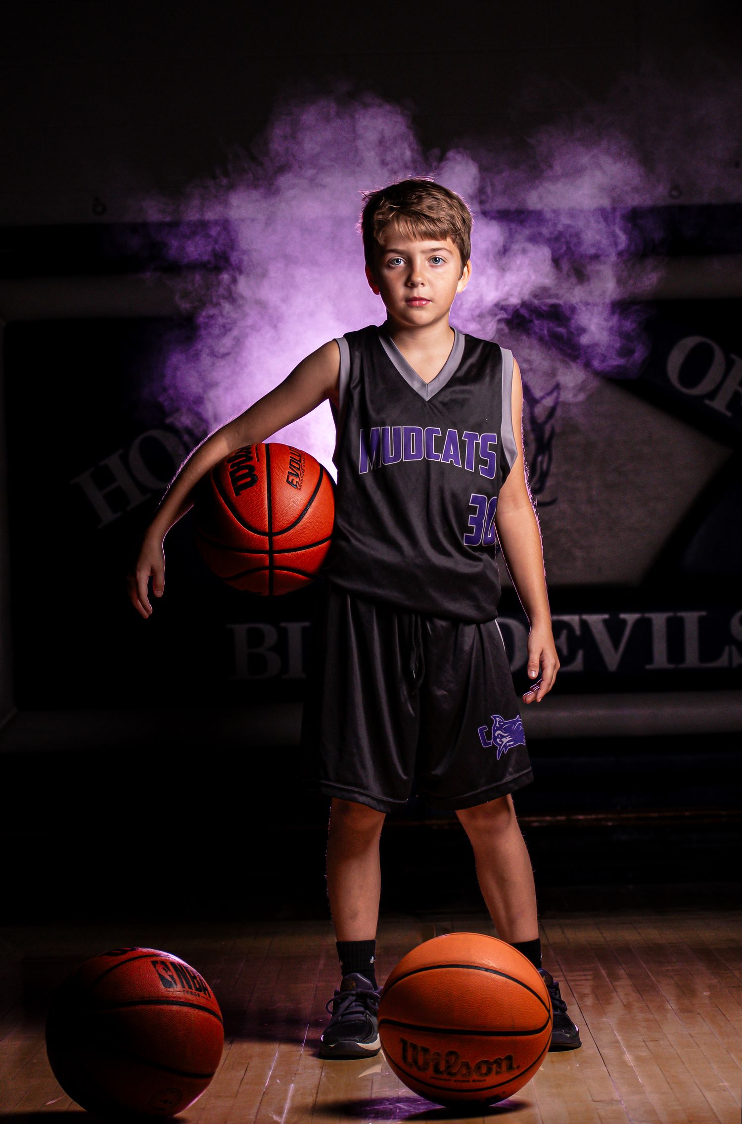Youth basketball player in black uniform poses with basketballs against purple smoke background.