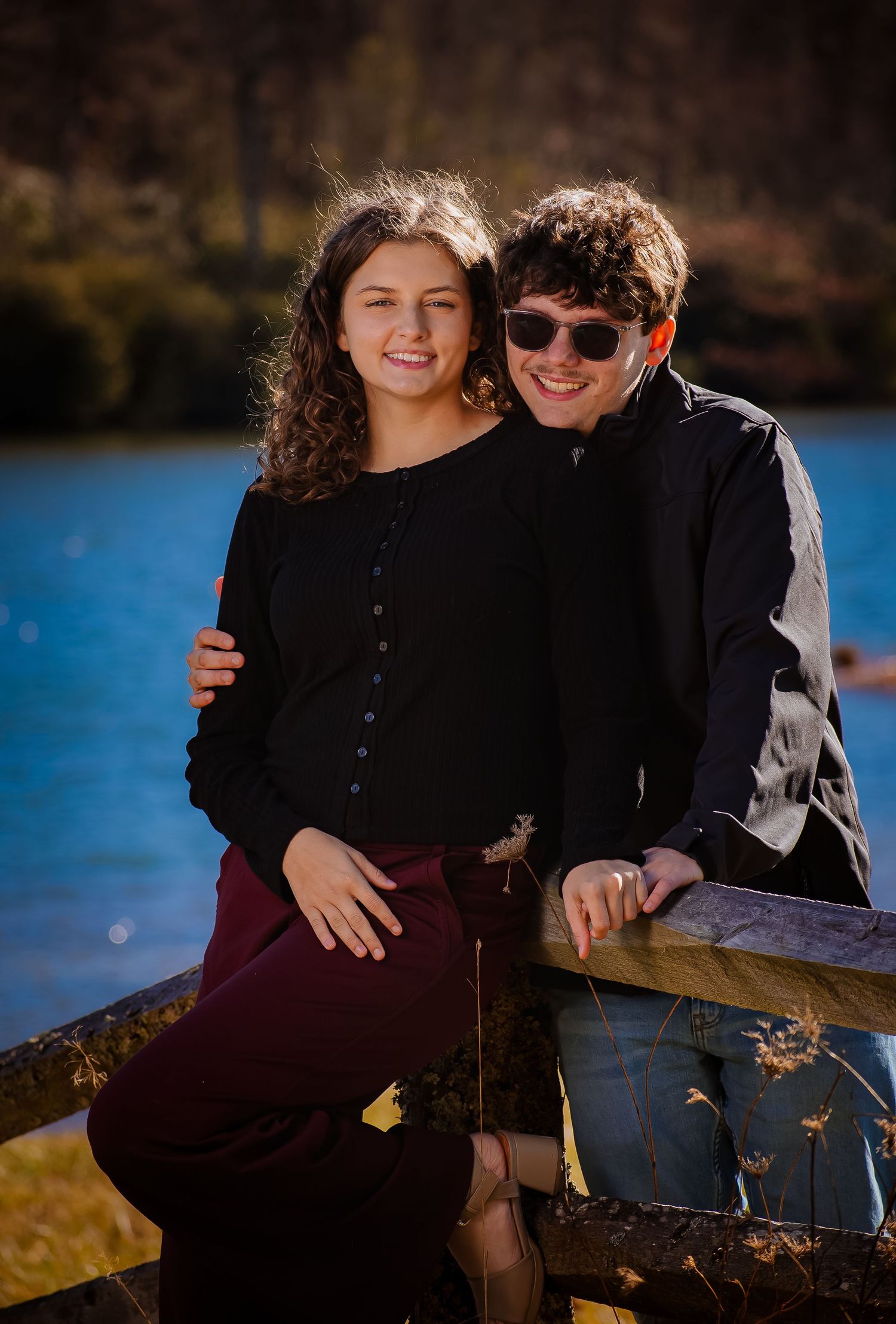A couple sitting together on a wooden fence by a lake on a bright autumn day.