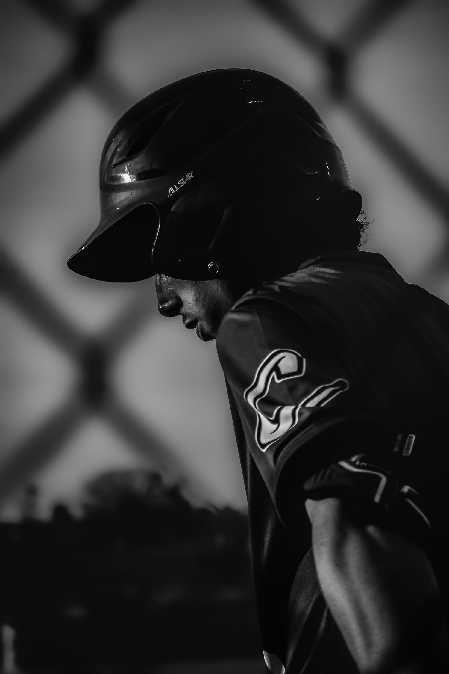 Black and white photo of a baseball player in helmet and jersey, standing near a chain-link fence.