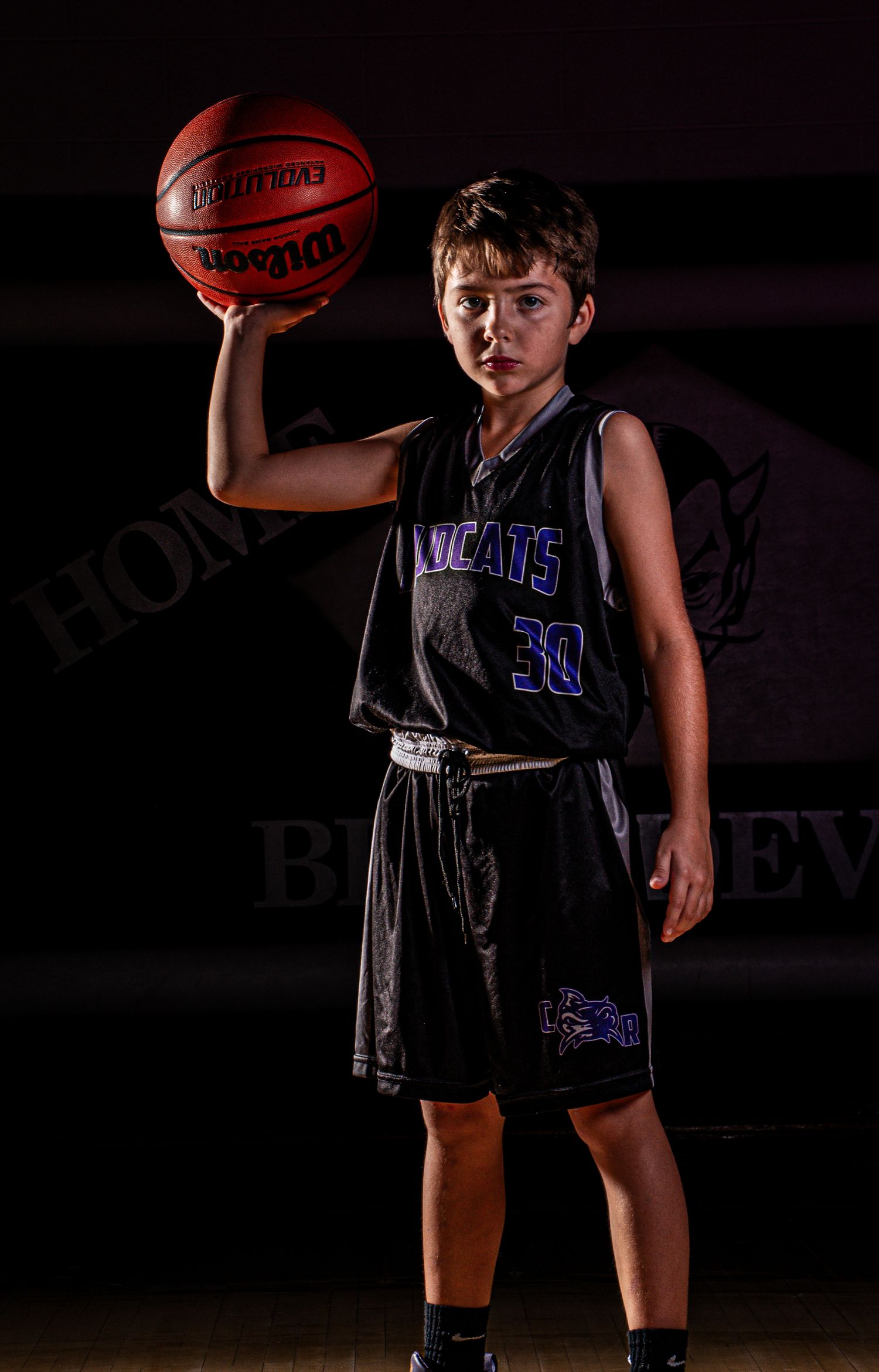 Young basketball player in black Wildcats uniform holds up a basketball in dramatic dark lighting.