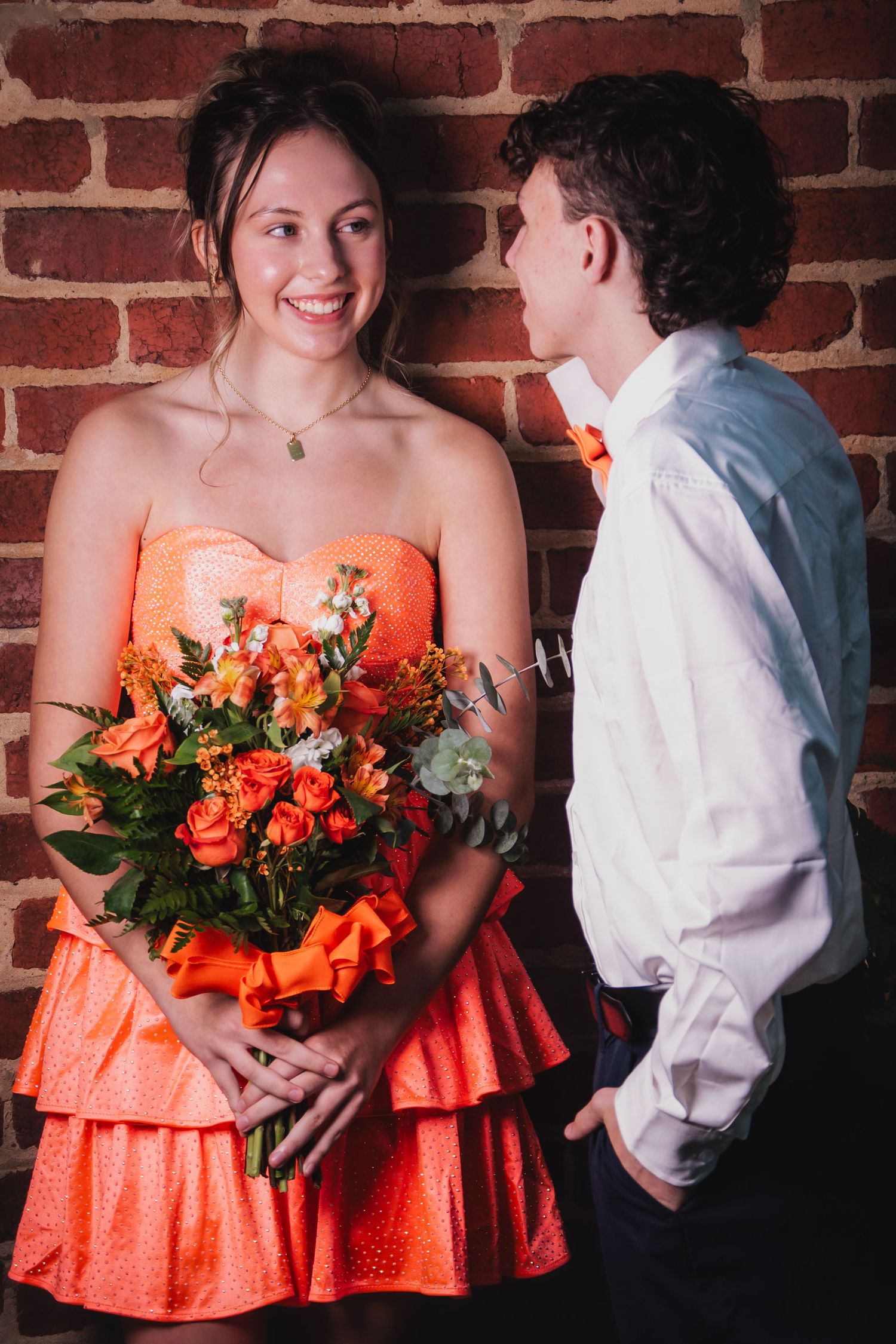 A couple in wedding attire share a moment against a brick wall, with the bridesmaid holding a vibrant orange bouquet.