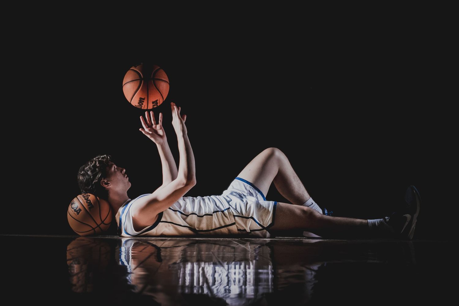 Basketball player reclines on court tossing ball in dramatic studio lighting.