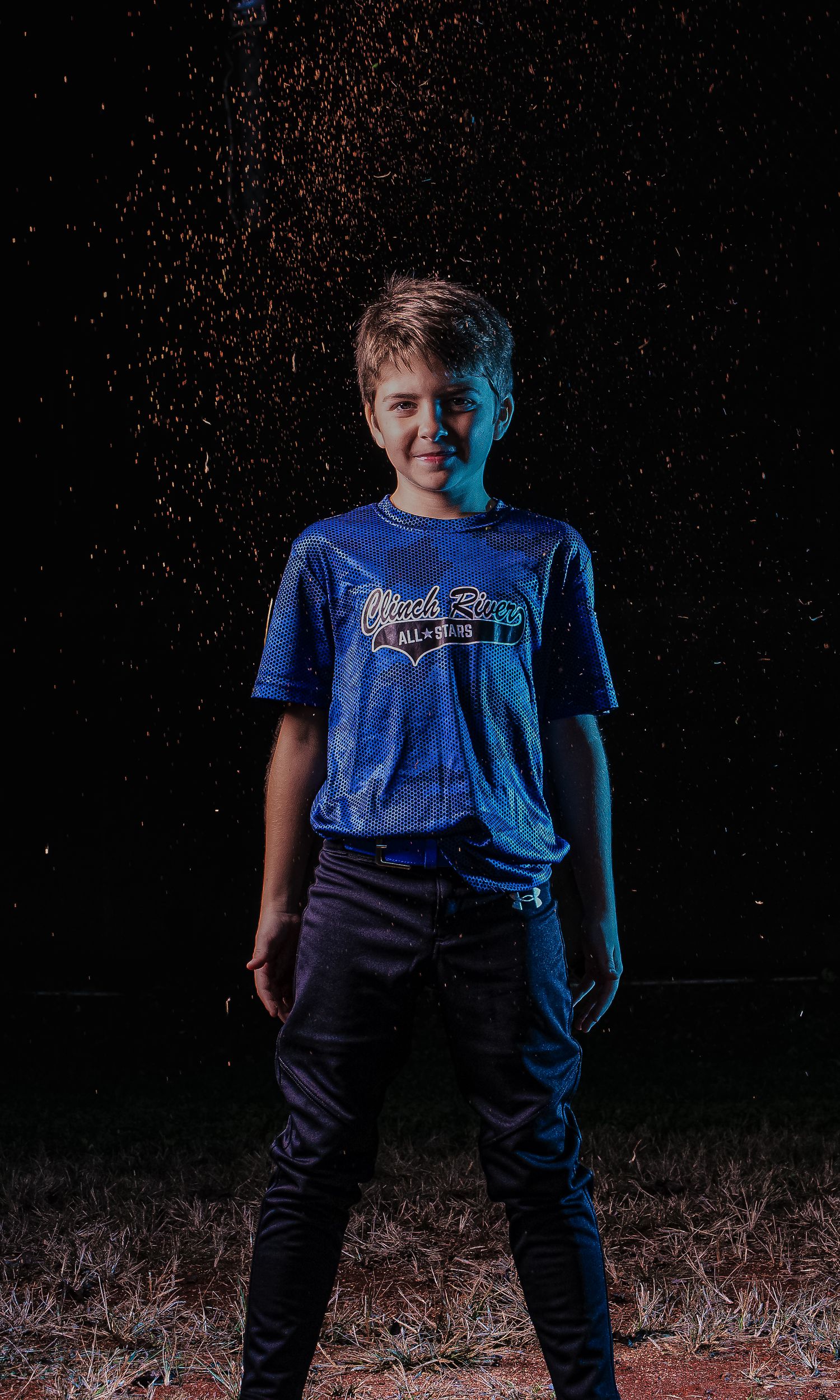 Youth athlete in blue baseball uniform poses against dark background.
