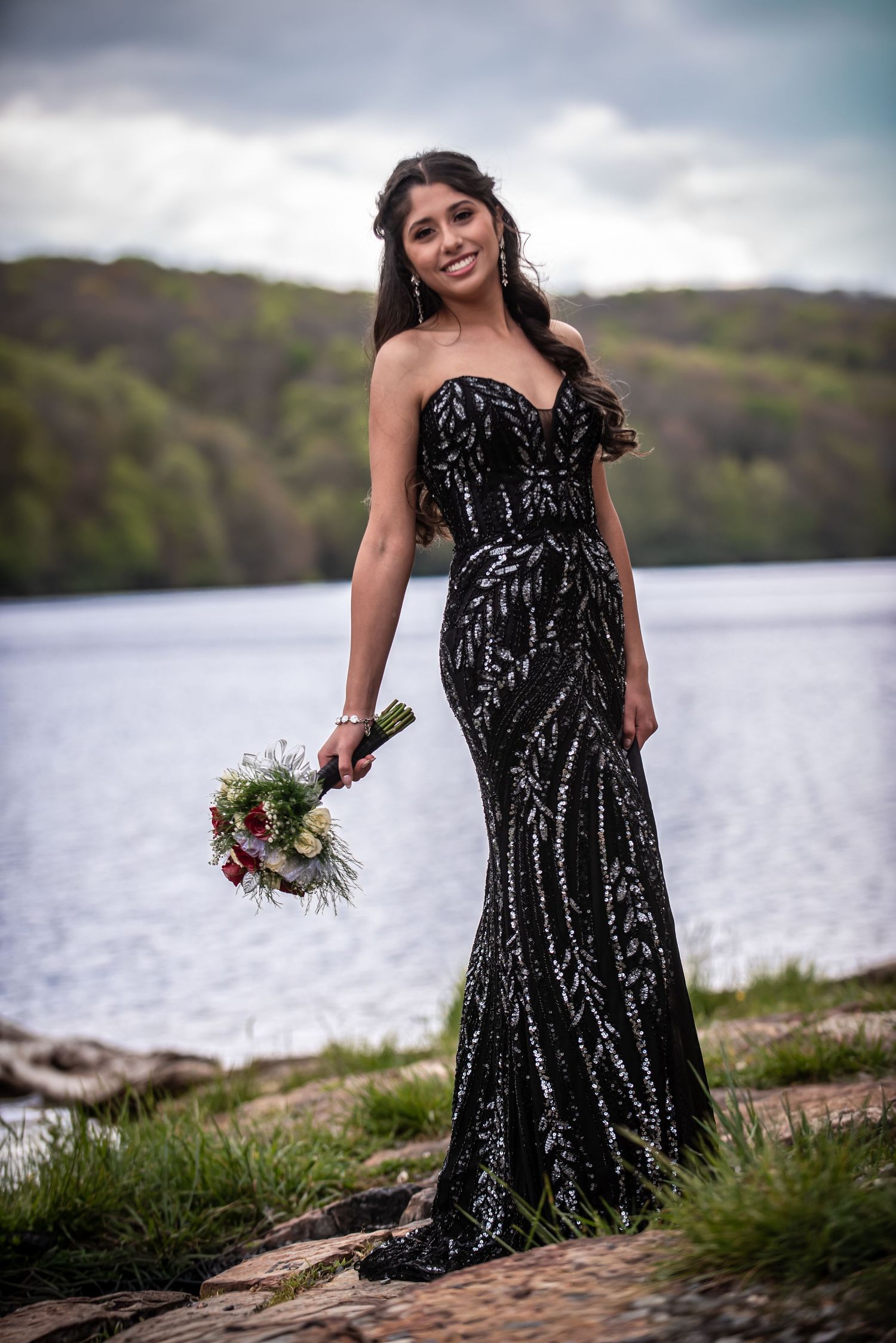 Smiling woman in black sequin strapless gown holding bouquet outdoors by lake.