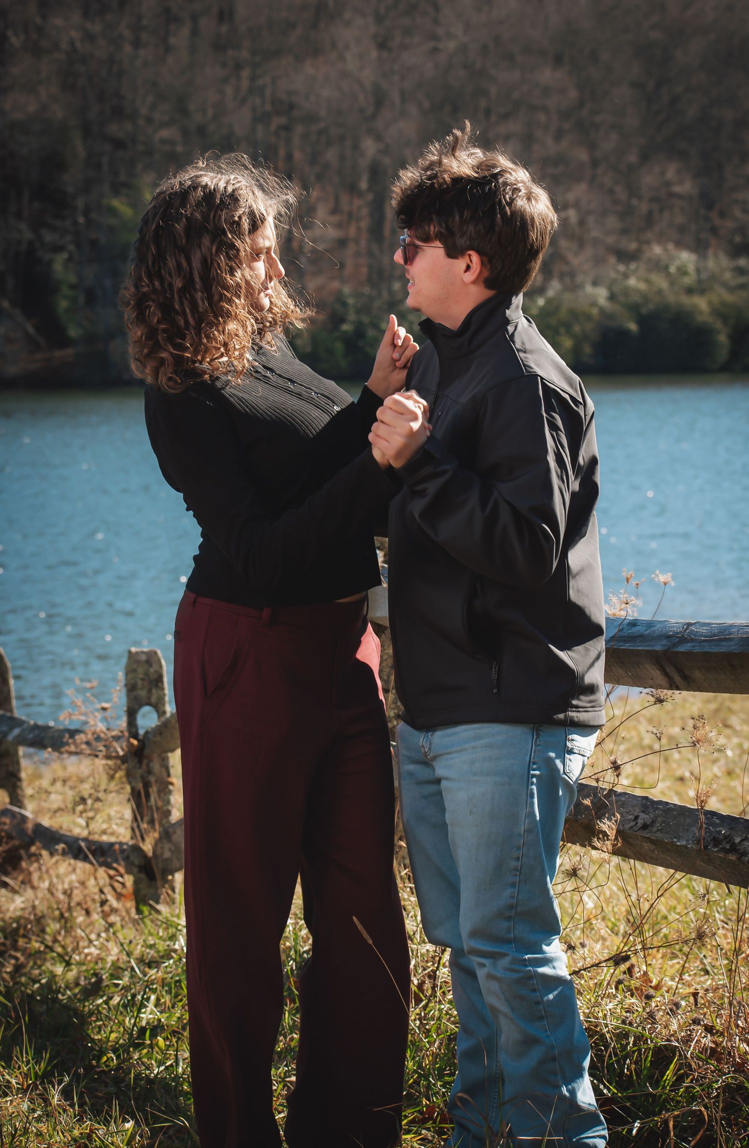 A couple dancing together by a scenic lake with a wooden fence in the background on a sunny day.