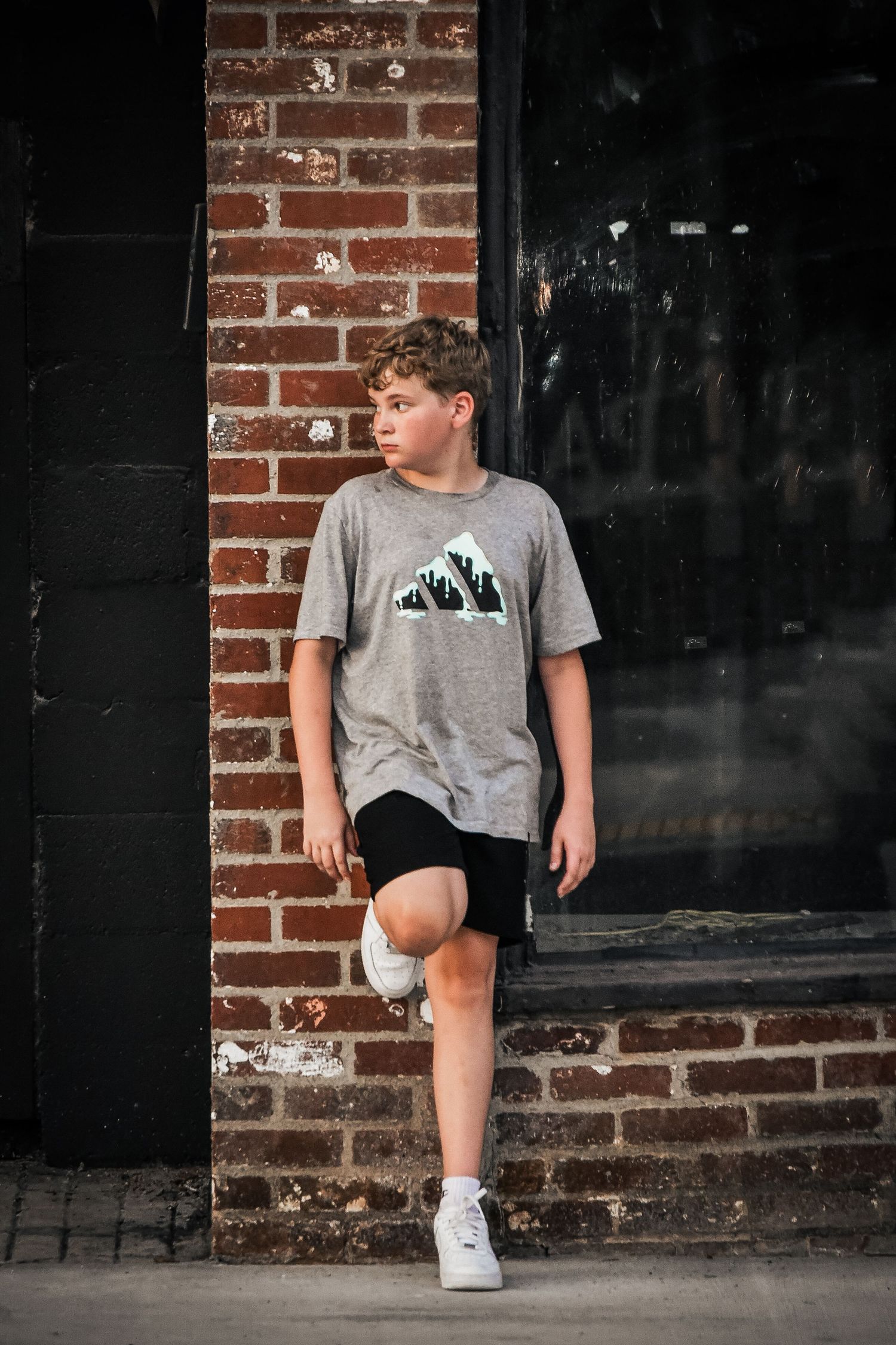 Person in grey t-shirt and black shorts leans against dark brick wall.