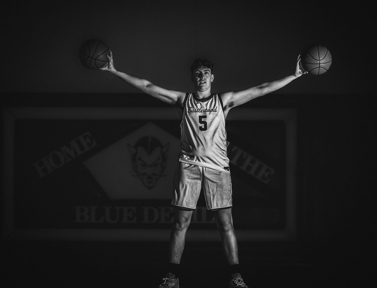 Basketball player spreads arms while holding basketballs in dramatic arena lighting.