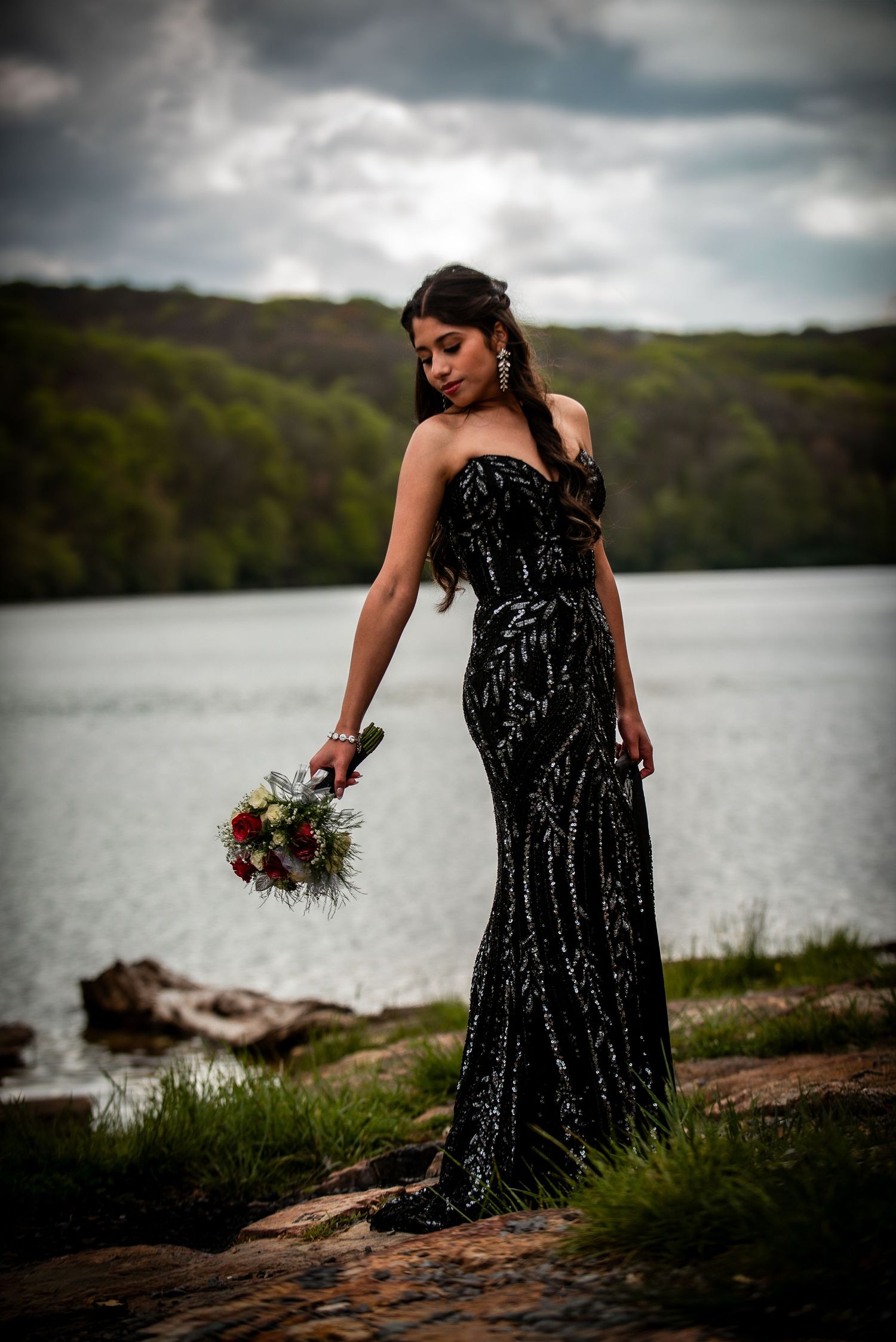 Woman in black sequin strapless gown holding bouquet by lakeside with lush green hills and dramatic cloudy sky.