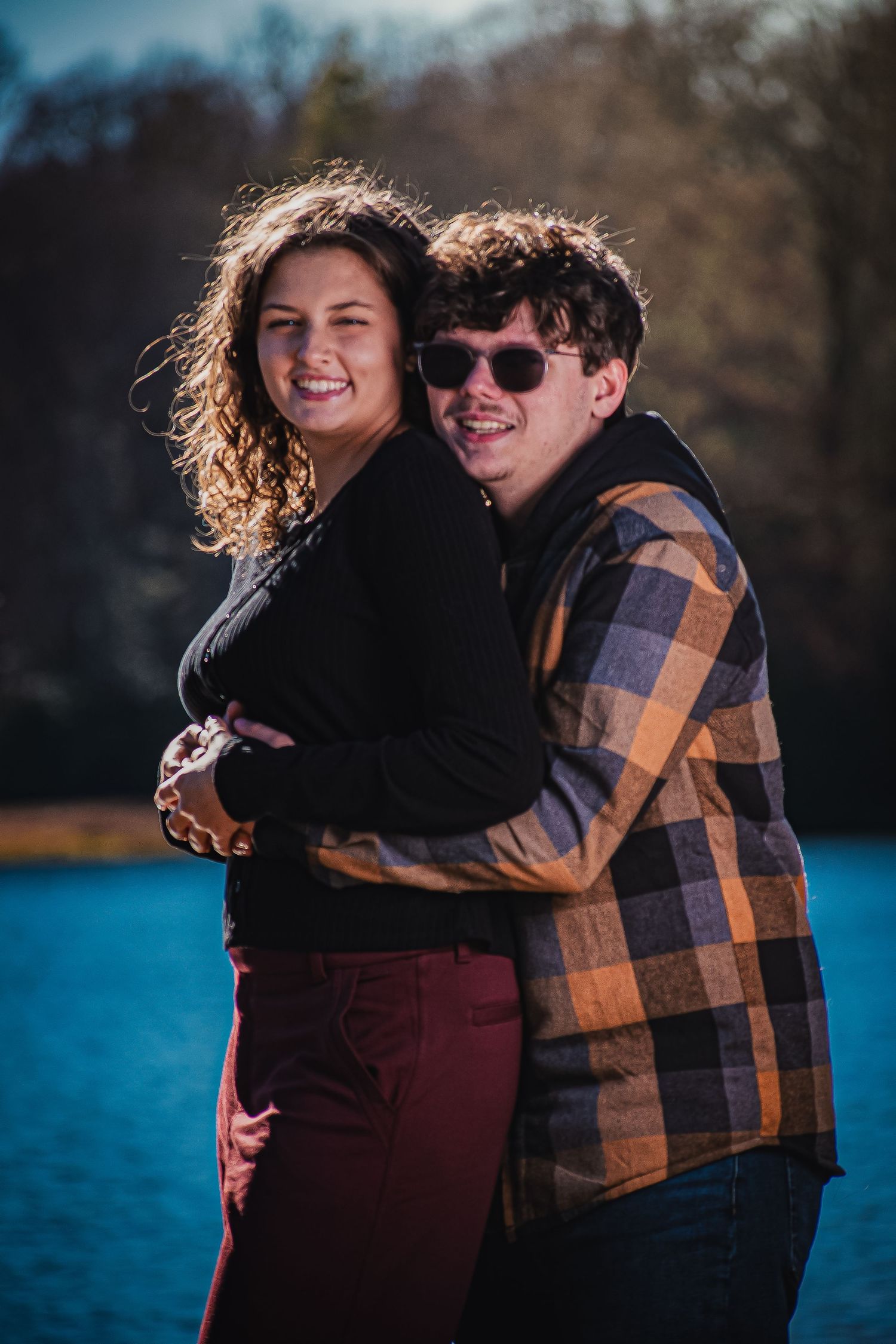 A couple shares a tender moment by a lake on a sunny day while wearing casual fall clothing.