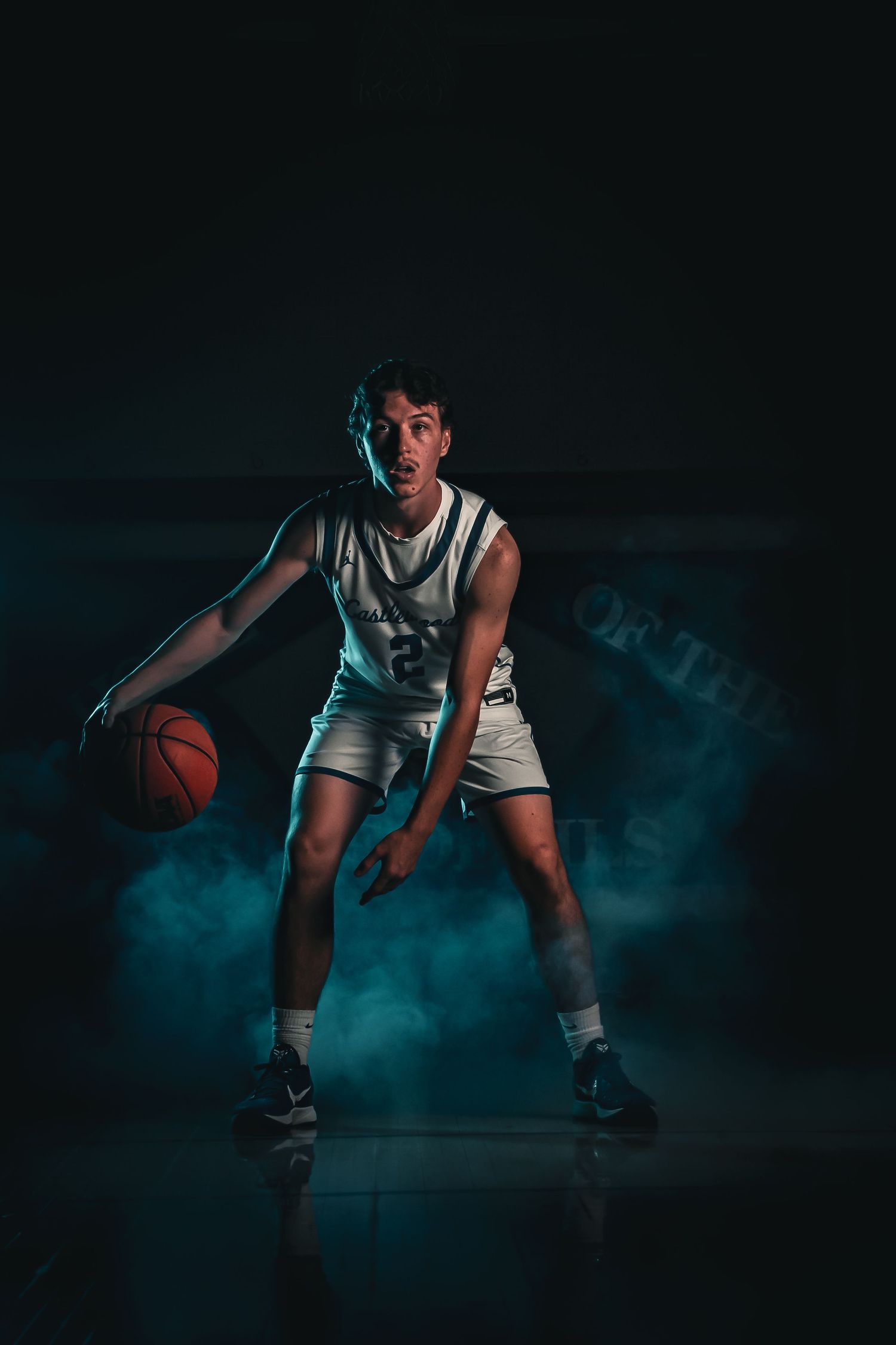 Basketball player in white uniform crouches in defensive stance against dramatic dark background with blue smoke effects.