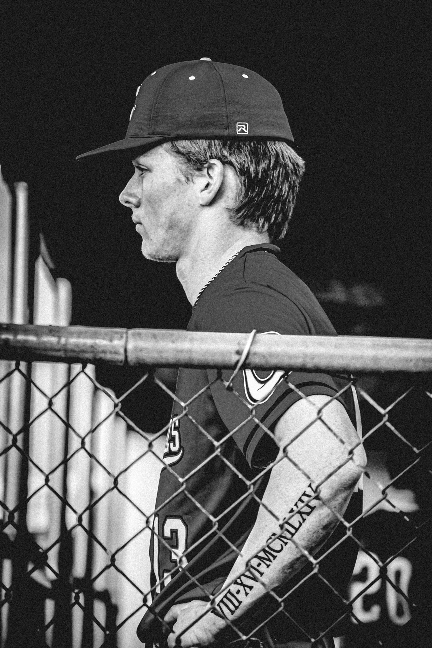 Black and white photo of a young baseball player in uniform leaning against a chain-link fence in a dugout.