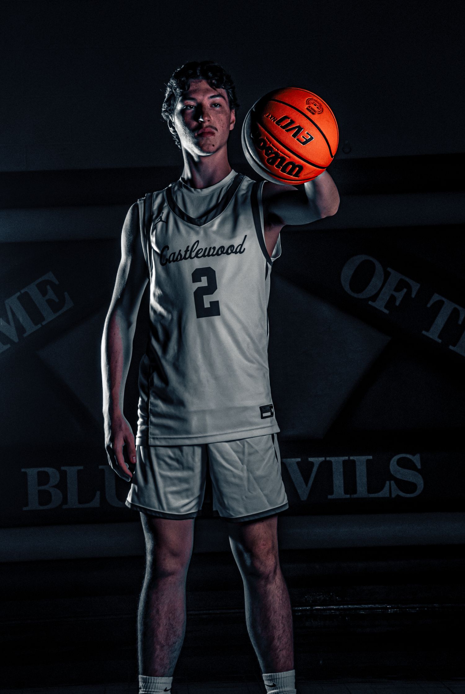 Basketball player in white uniform holds ball while standing in dark studio with team logo visible.