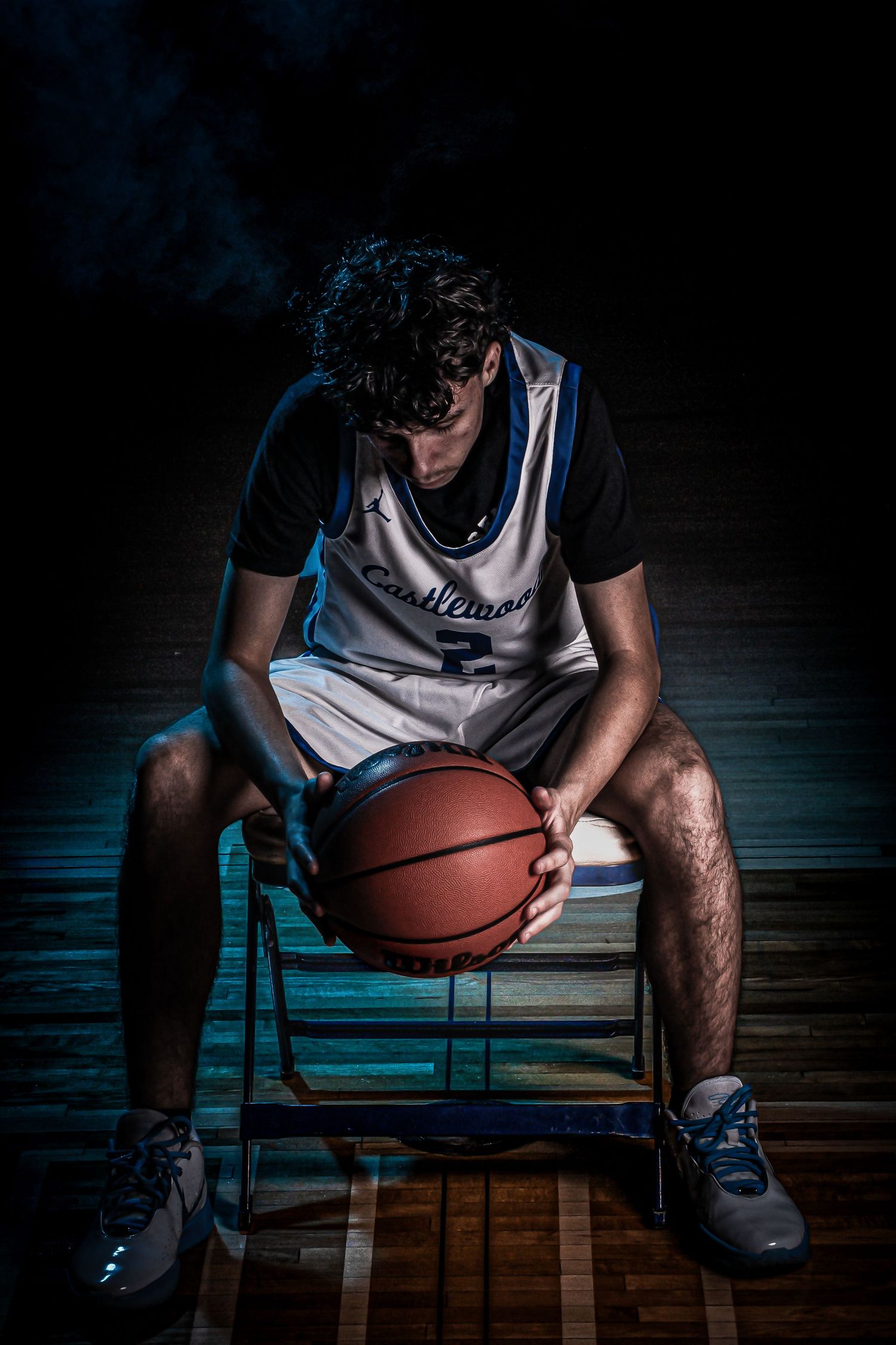 Dramatic low-key portrait of a basketball player sitting on bench holding ball in dark gym.