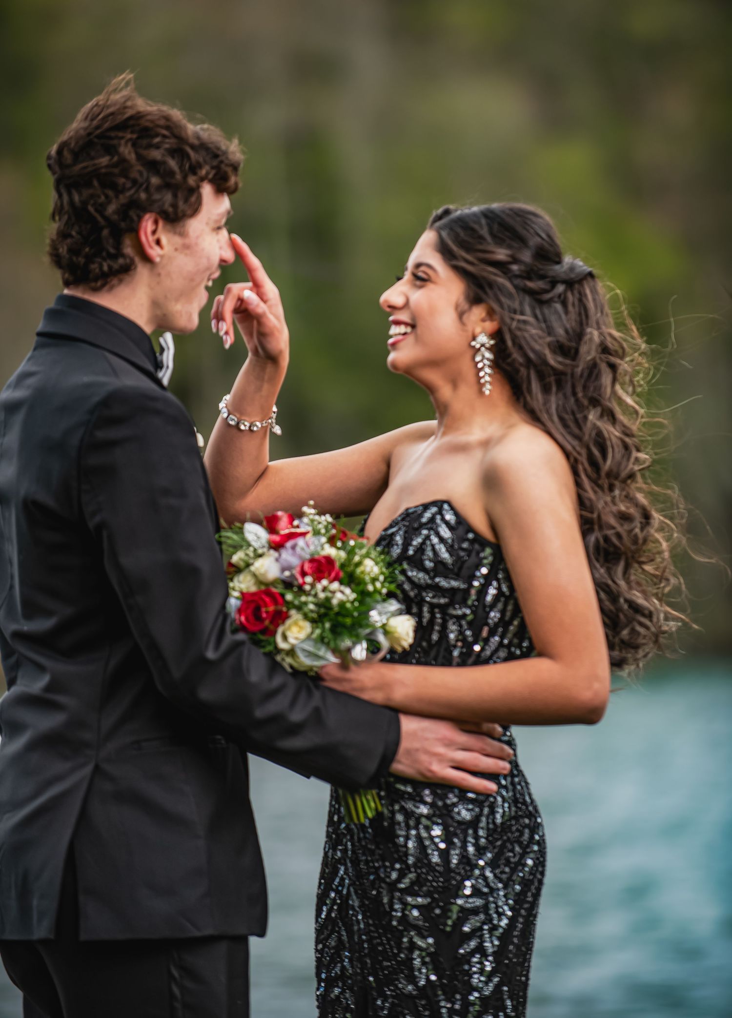 Couple in formal prom attire smiling, girl in black sequin gown holding red roses bouquet, boy in black suit.