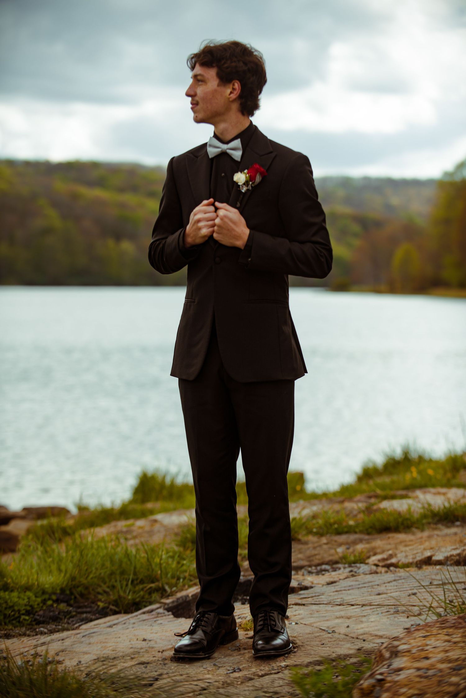 Groom in black tuxedo with boutonniere stands on rocky shore by lake, lush green hills in background.