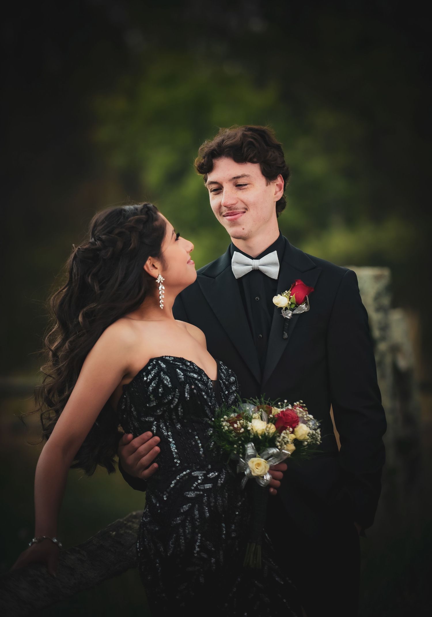 A couple in formal prom attire, the girl in a black sequin gown and the boy in a tuxedo with boutonniere, share a romantic moment.