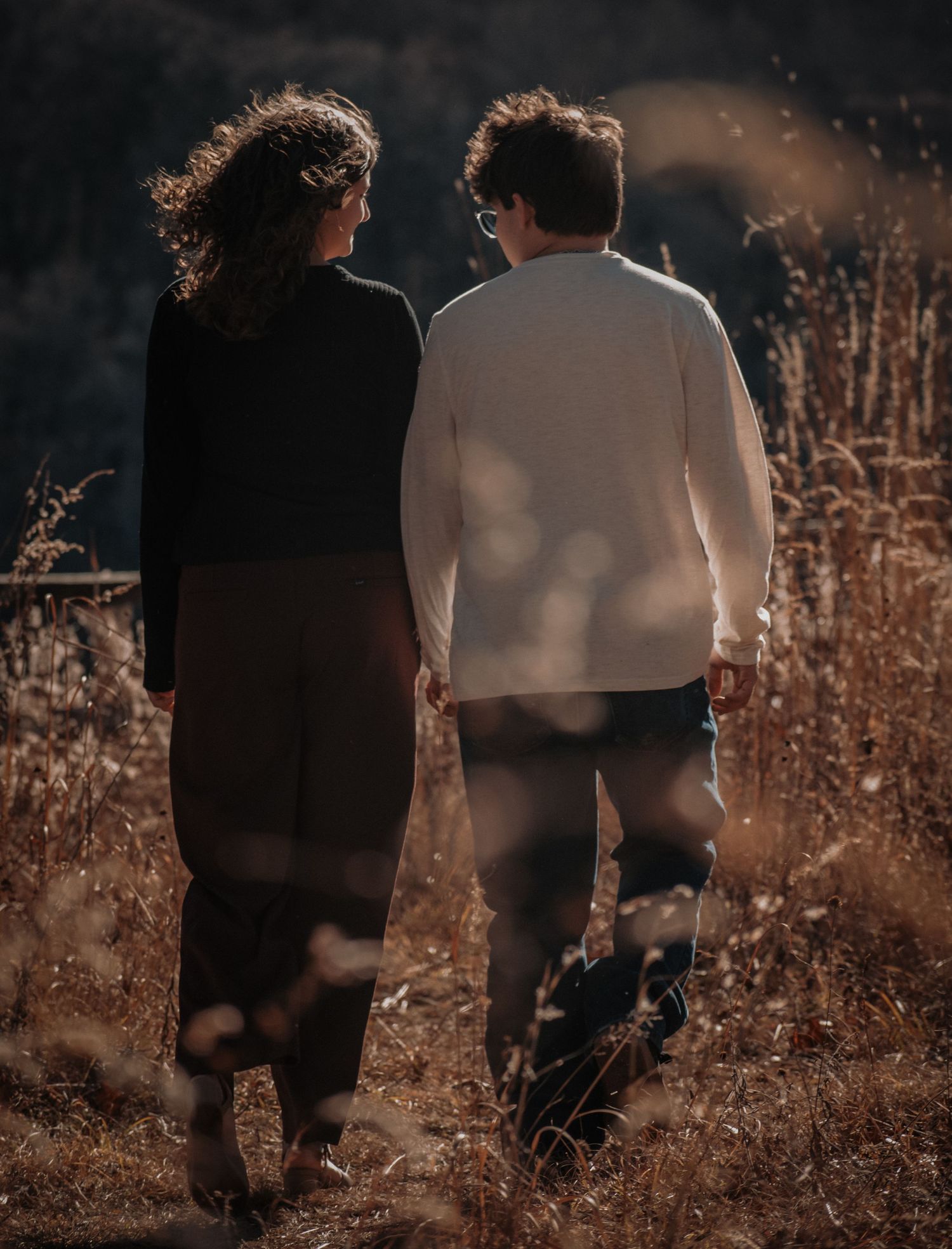 A couple walks together through tall golden grass at sunset.