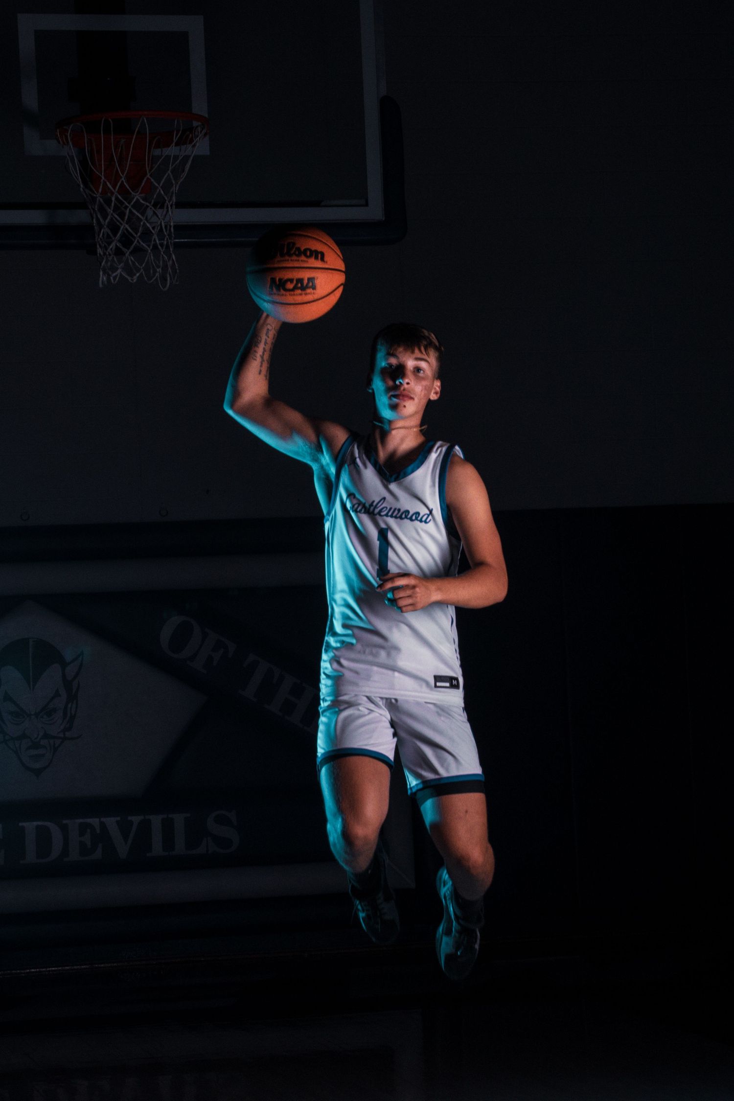 Basketball player jumps with ball in dramatic lighting against dark gym background.