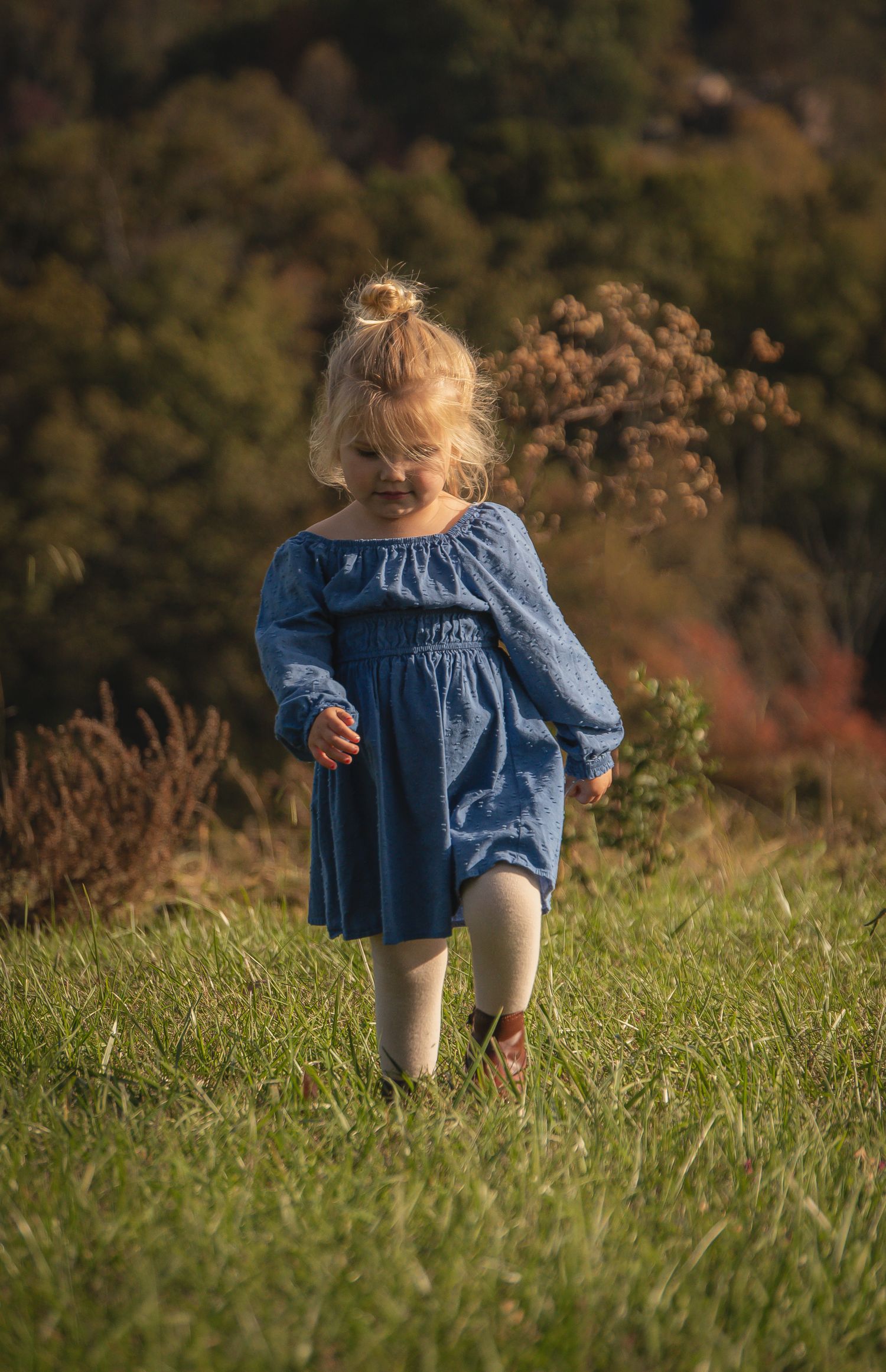 Child in denim dress explores grassy hillside during autumn afternoon.
