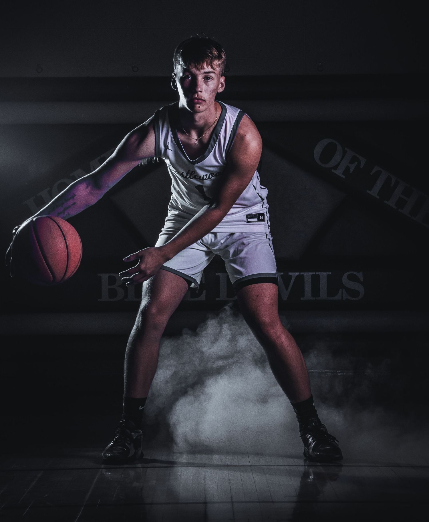 Basketball player in white uniform crouches with smoke effect on dark court.
