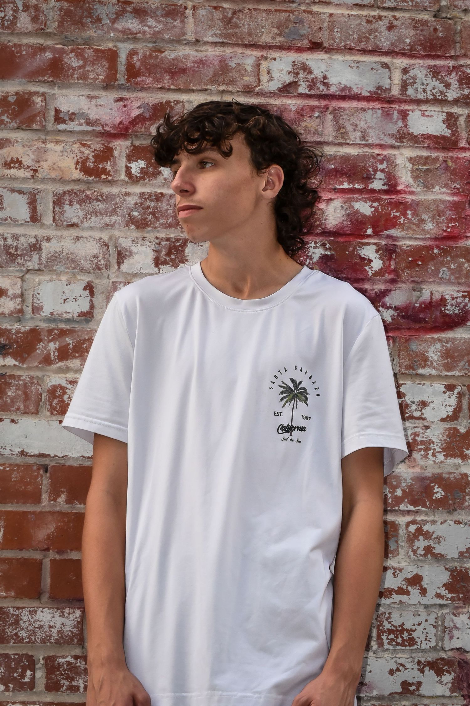 Person in white t-shirt with small palm tree logo stands against rustic brick background.