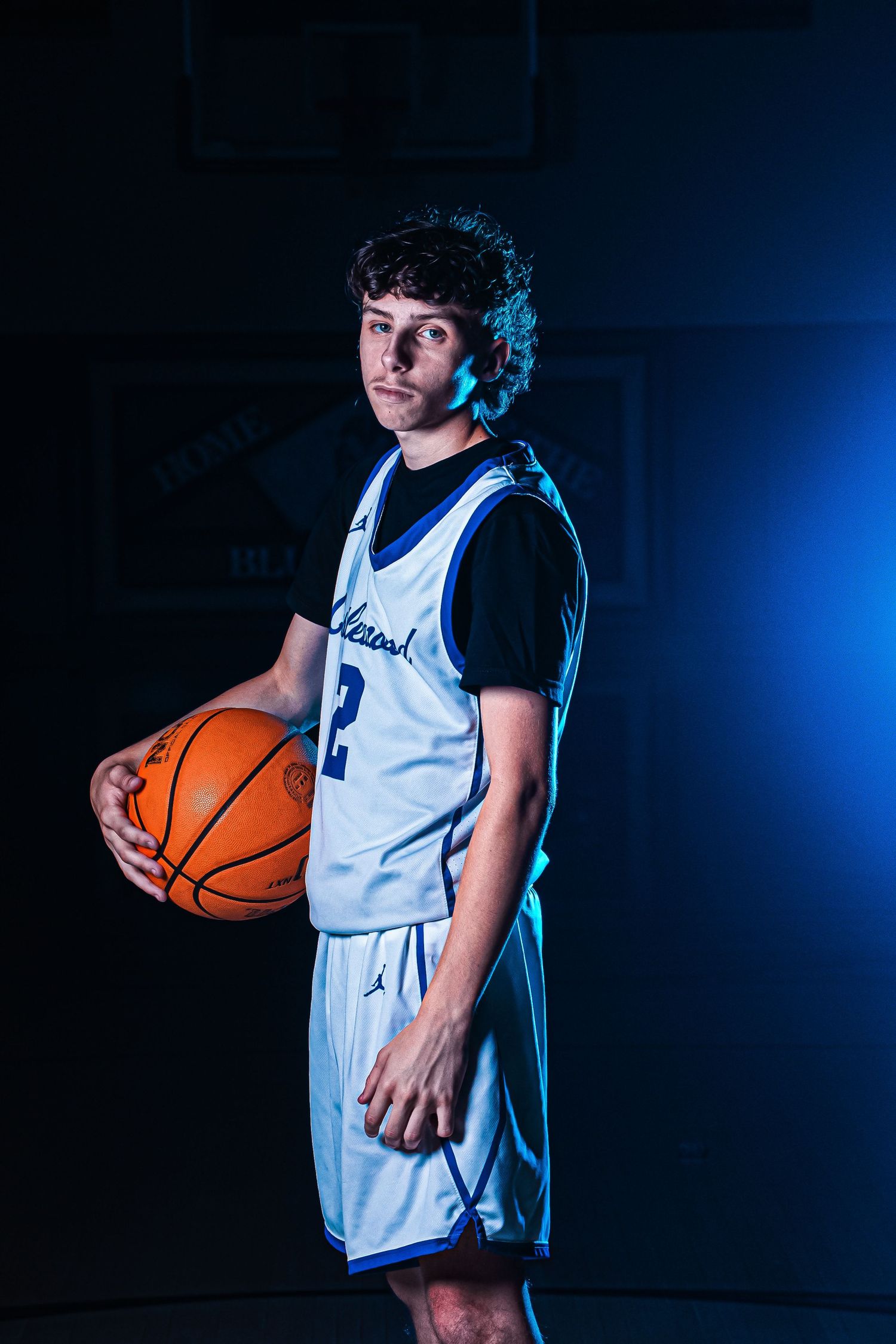 Basketball player in white and blue uniform number 2 poses with ball against dark background with dramatic lighting.