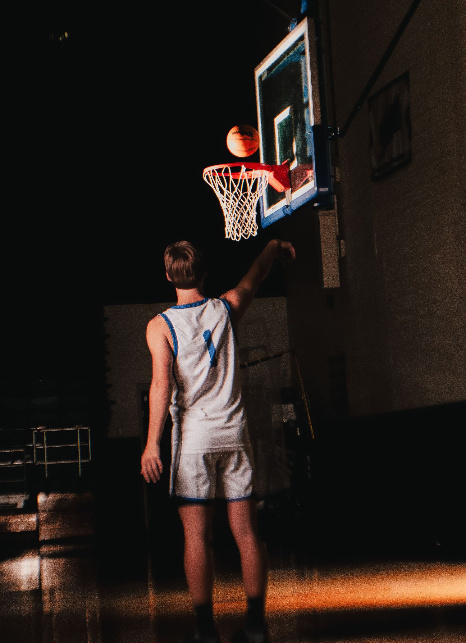 Basketball player stands before hoop in dimly lit gymnasium.